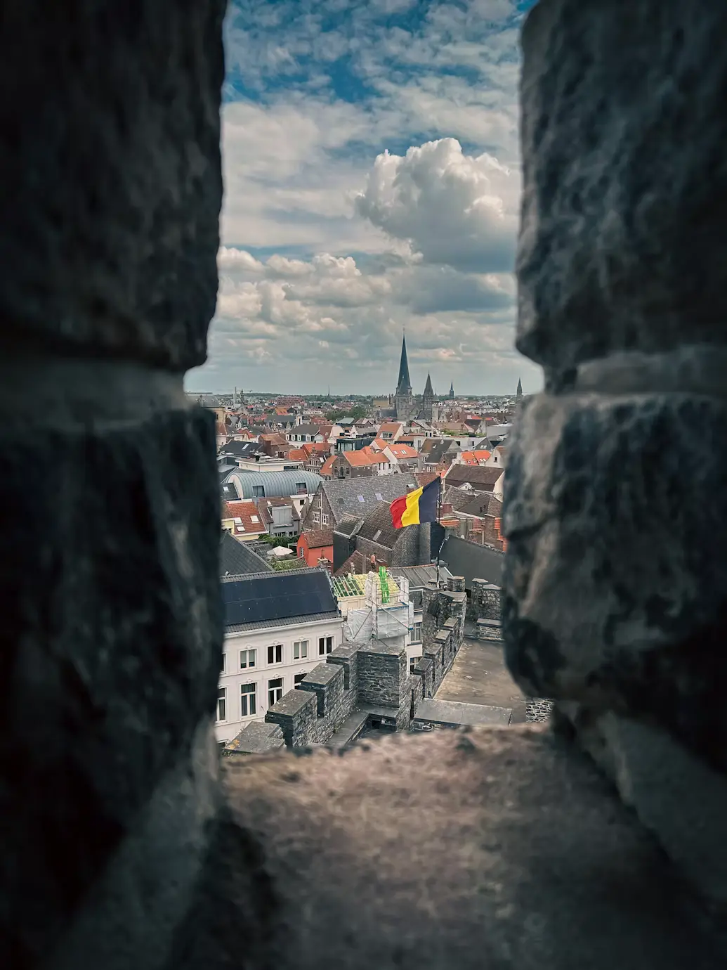 View through a castle battlement opening over Ghent's rooftops, with a church spire and Belgian flag in the distance.