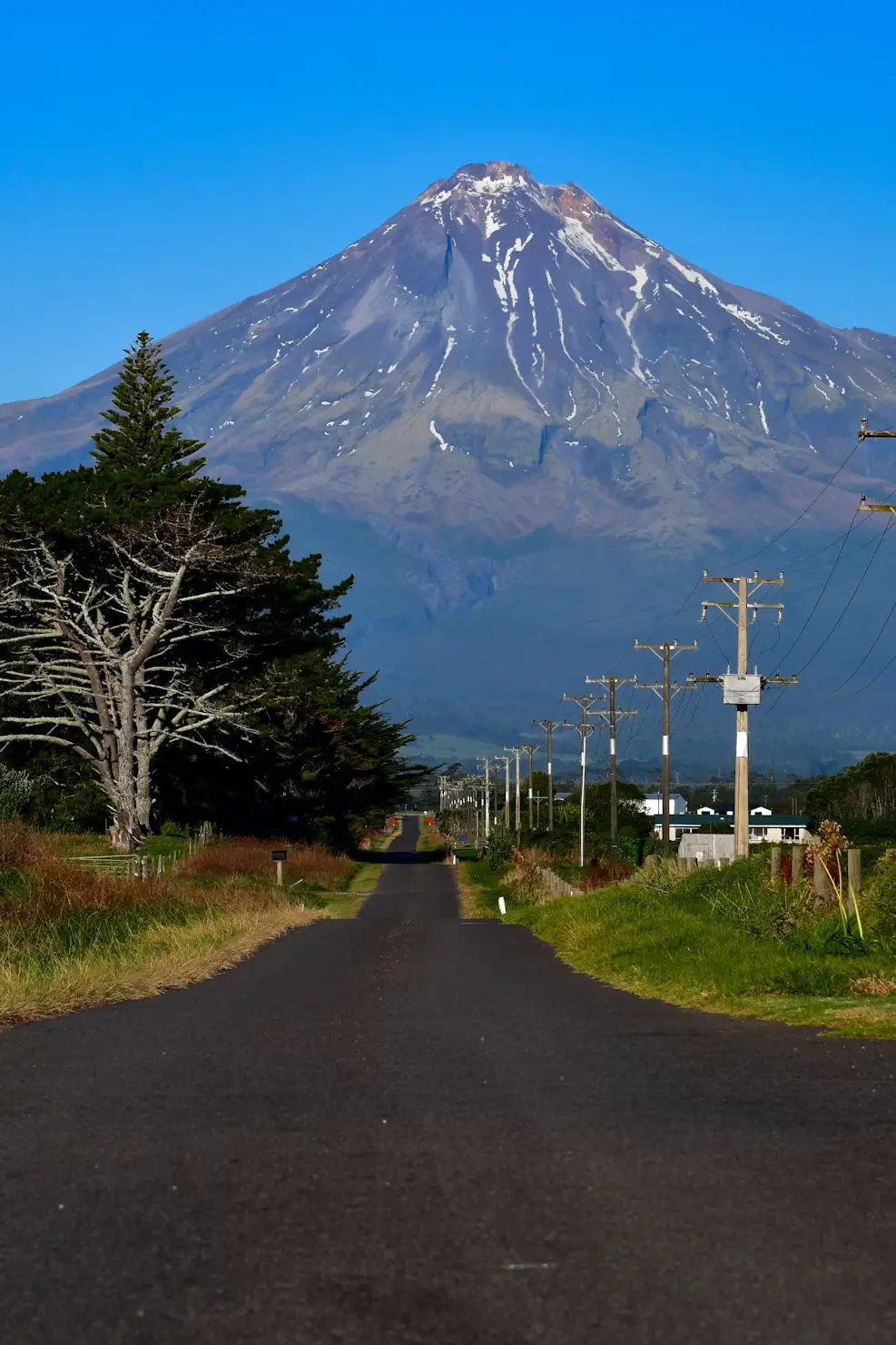 Snow-streaked volcanic mountain rising above a rural road lined with power poles.