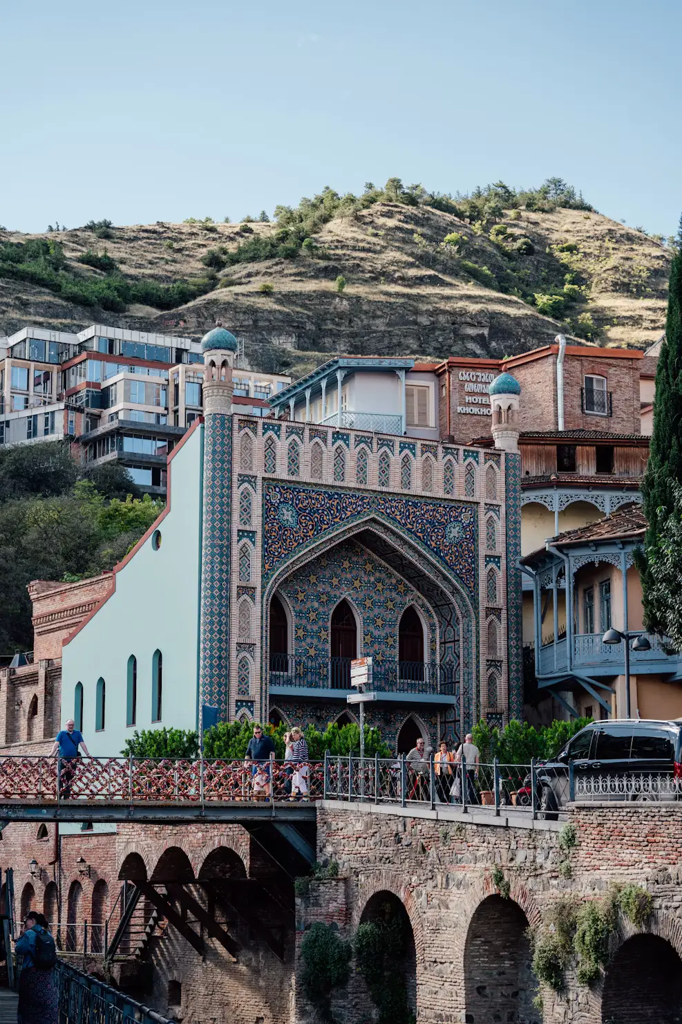 Ornate blue-tiled historic building beside a bridge with hillside homes and dry hills in the background.