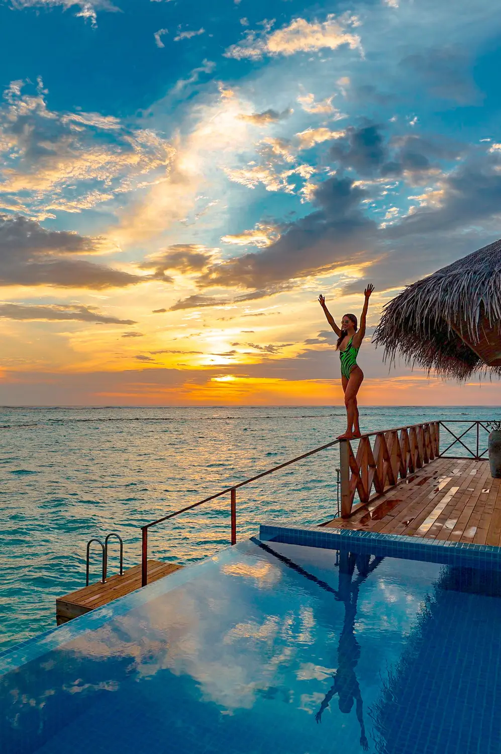 Person standing with arms raised on overwater villa deck with infinity pool at golden sunset over Maldives ocean horizon