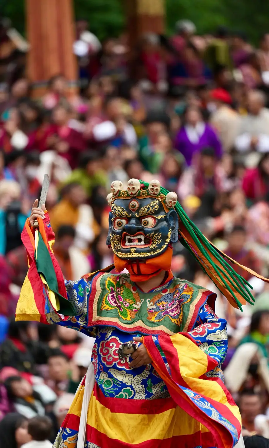 Masked dancer in a colorful silk costume performing a traditional Cham dance before a crowd at a festival in Bhutan.