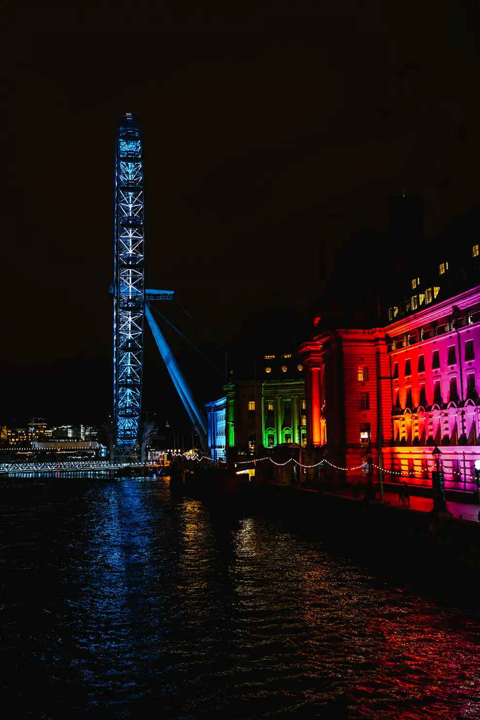 Night view of the London Eye glowing blue beside buildings lit in rainbow colours, their reflections shimmering on the Thames.