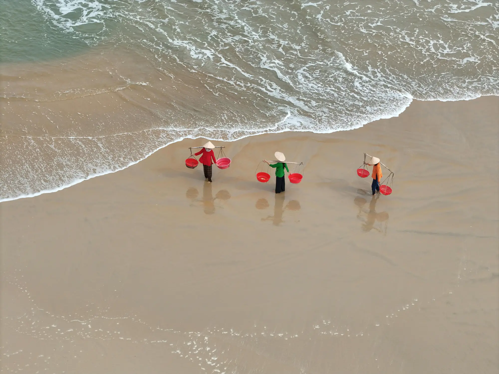 Three Vietnamese women wearing traditional nón lá conical hats carrying red baskets on shoulder poles along a sandy beach shoreline, aerial drone view, Vietnam