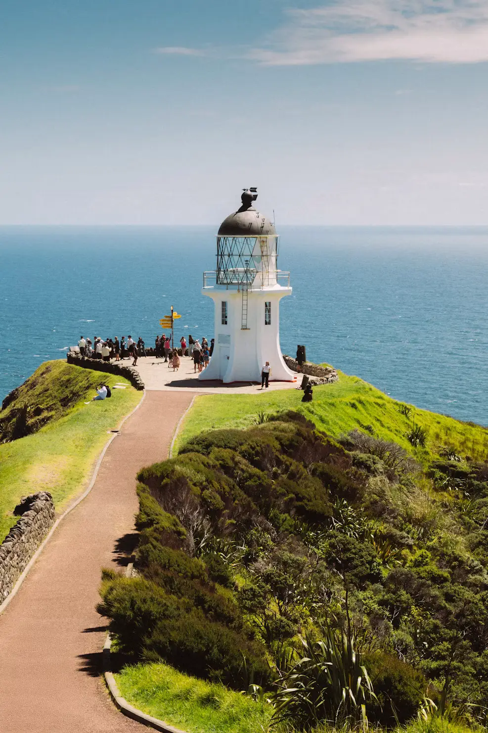White lighthouse on a grassy headland overlooking the blue ocean with visitors nearby.