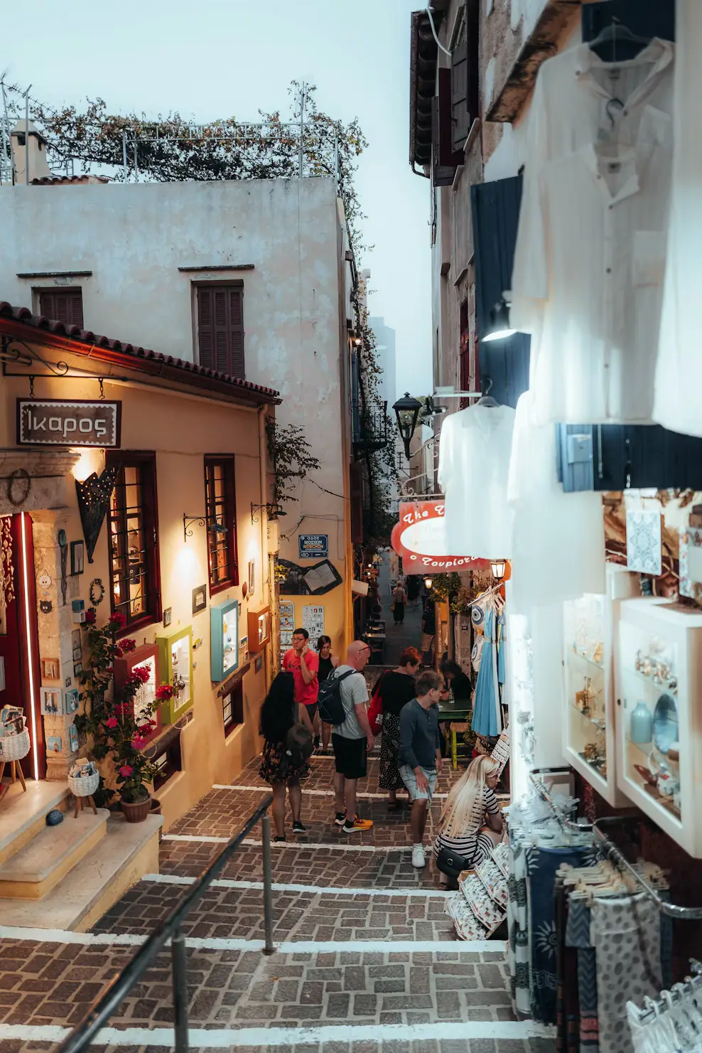 A narrow pedestrian shopping street in a Greek town, lined with shops and strolling visitors.
