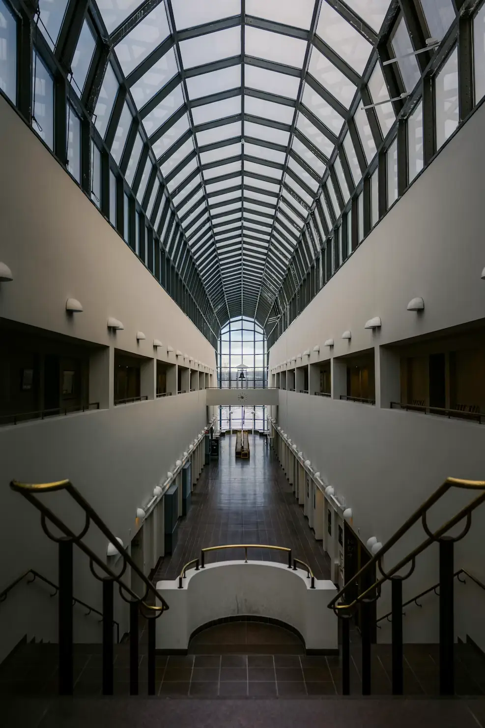 Modern building atrium with a vaulted glass ceiling, symmetrical balconies, and a central staircase.