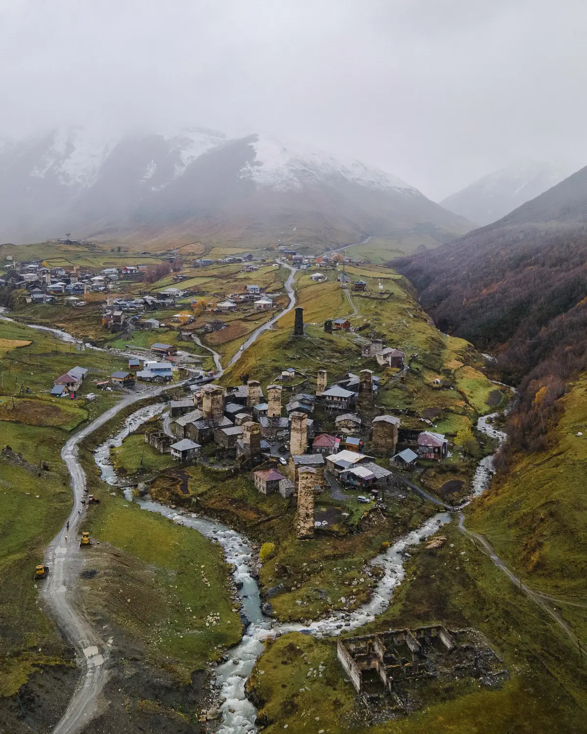 Aerial view of a remote mountain village in a misty valley, with clusters of traditional stone tower houses, winding roads, and streams surrounded by green hills and snow-dusted peaks.