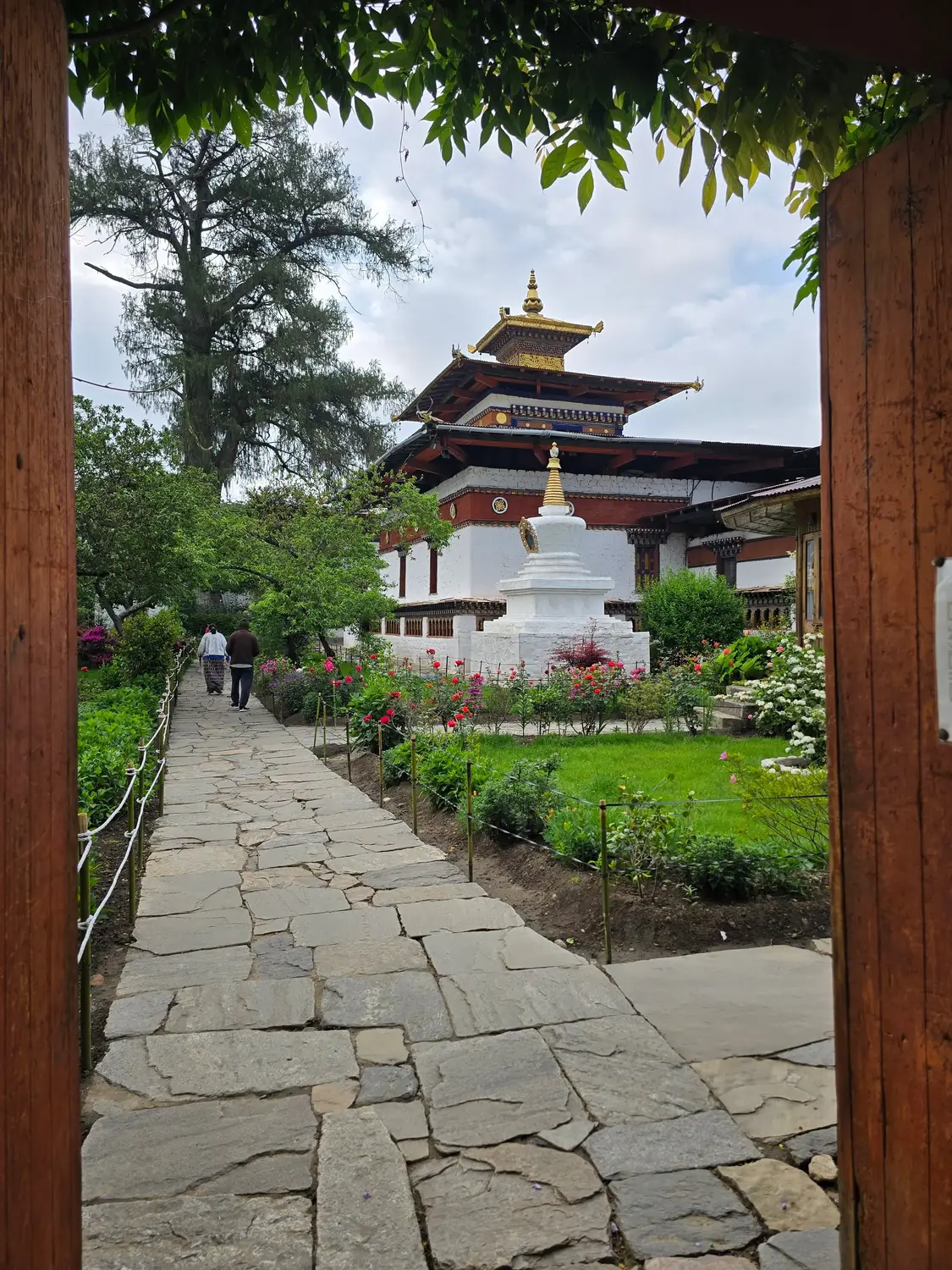 View through a wooden temple gateway of a white Buddhist stupa and traditional tiered temple, framed by gardens and a stone pathway in Bhutan.