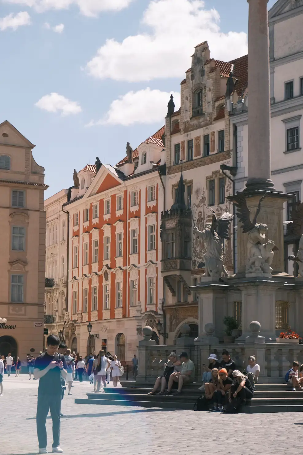 Person walking through a Prague square past a baroque fountain and colorful historic buildings