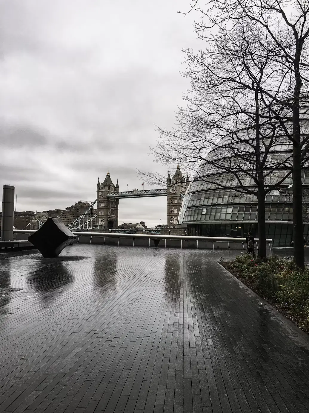 A rain-slicked riverside plaza with Tower Bridge and City Hall's glass facade in the background under an overcast sky.
