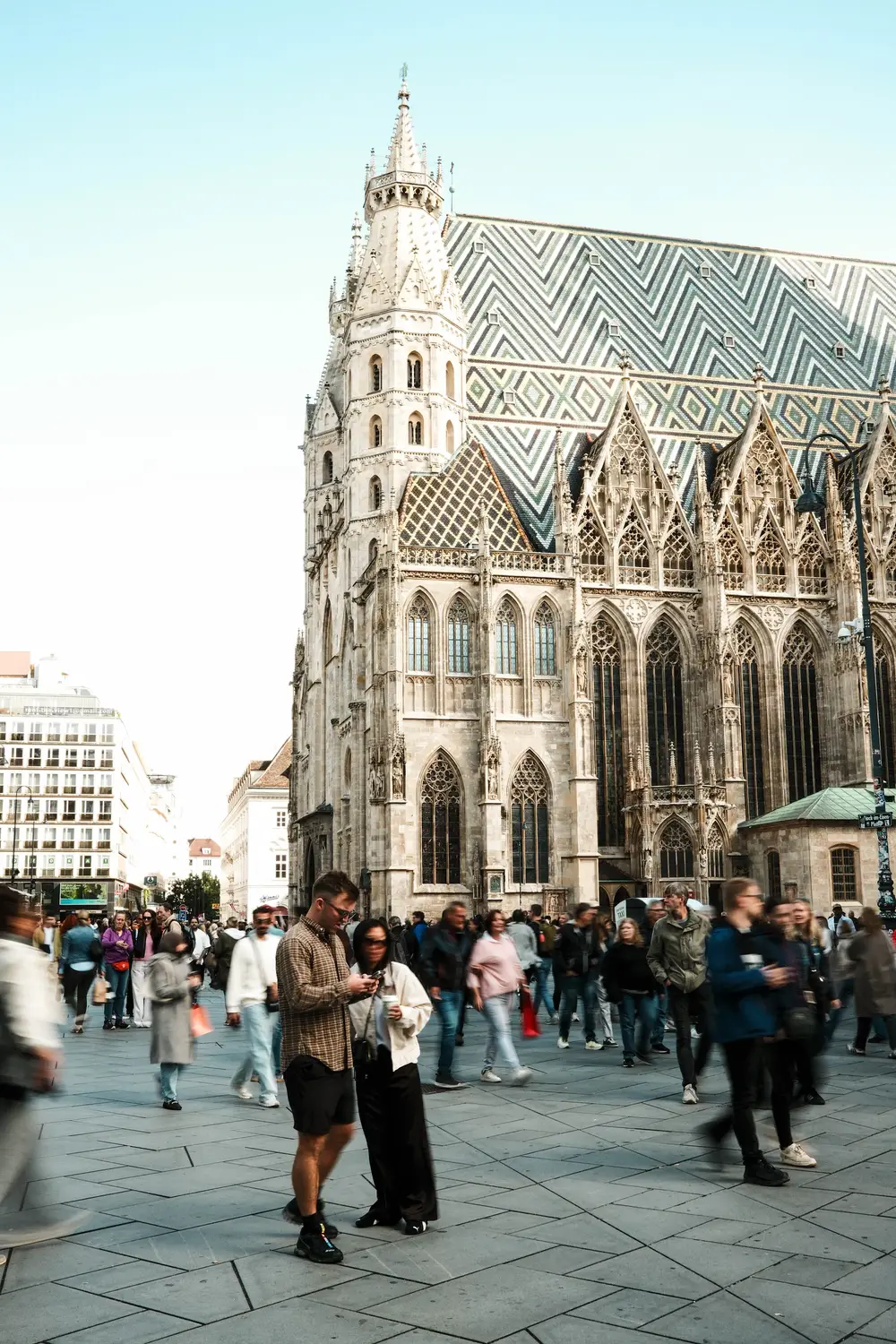 Pedestrians walking across a cobblestone square in front of a tall Gothic cathedral with an intricately patterned tiled roof.