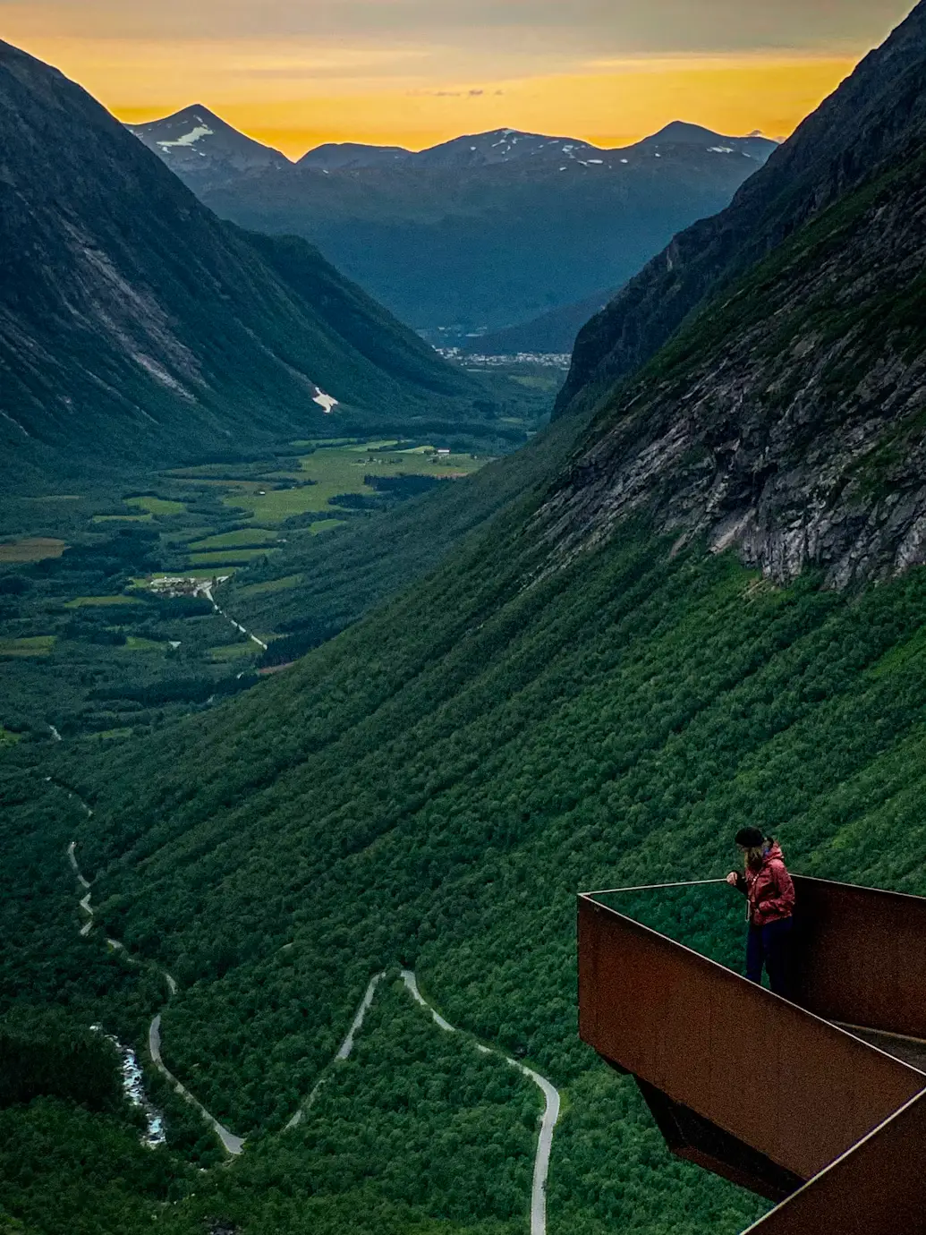 A person at a viewpoint overlooking a deep green valley with a winding road, mountains glowing under a golden sky.