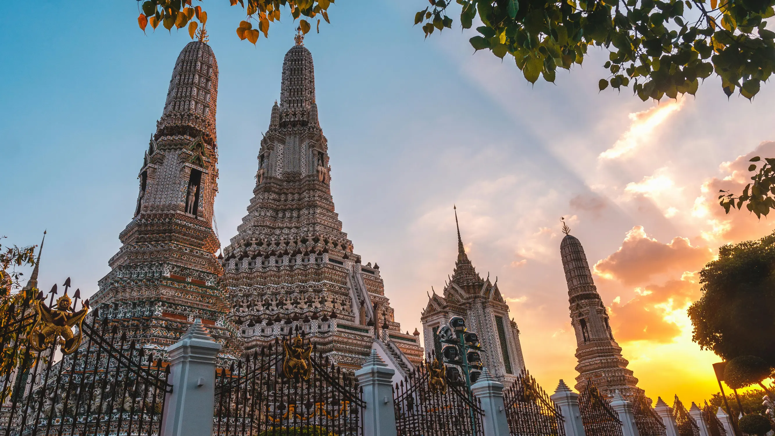 Low-angle view of Wat Arun's intricately decorated porcelain mosaic prangs and surrounding spires framed by tree branches against a dramatic golden sunset sky behind ornate iron gates, Bangkok, Thailand