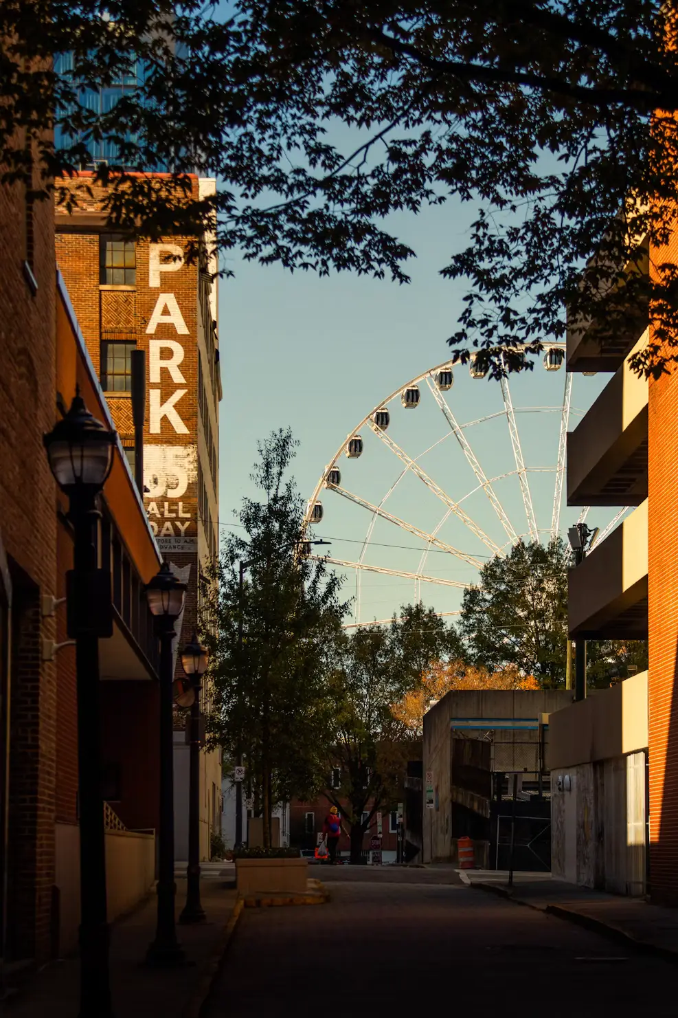 A city street with a large Ferris wheel visible between brick buildings and leafy trees.