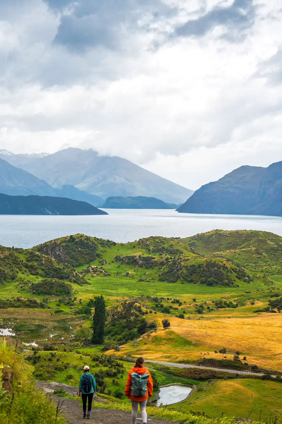 Two hikers overlooking rolling green hills, a lake, and layered mountains under a cloudy sky.