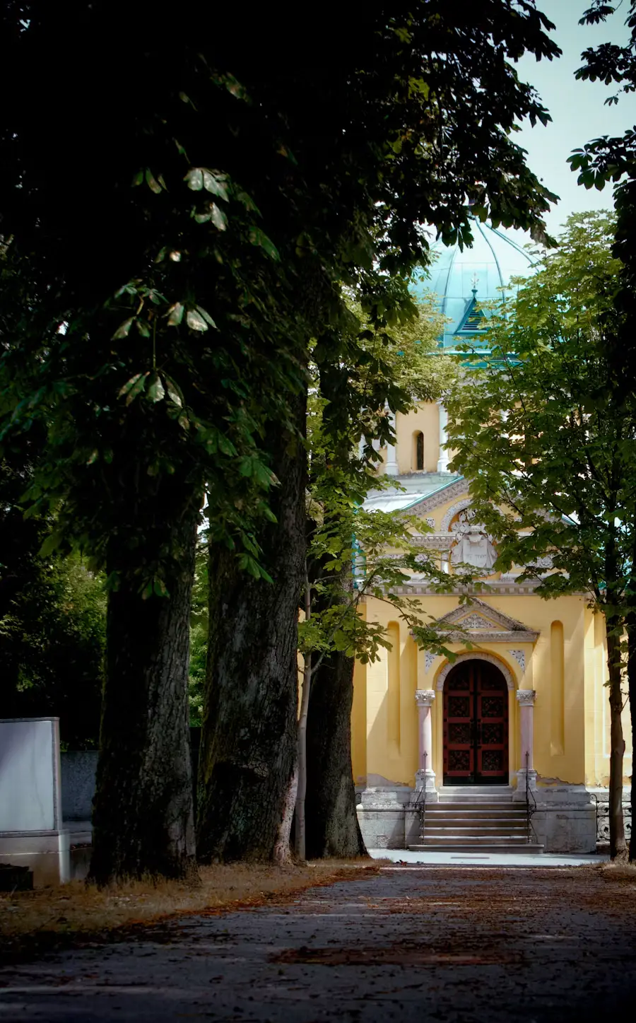 Small yellow chapel at the end of a tree-lined path dappled with sunlight.