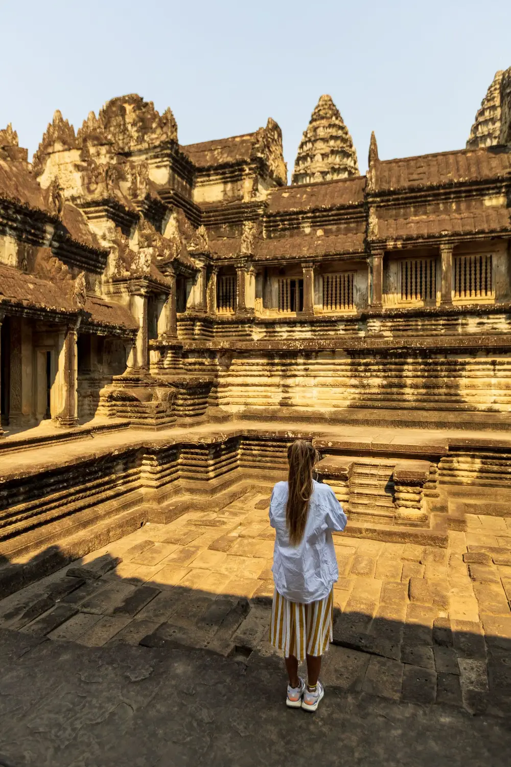 Female tourist standing facing ancient Angkor Wat stone temple towers and galleries glowing in golden morning sunlight, Siem Reap, Cambodia