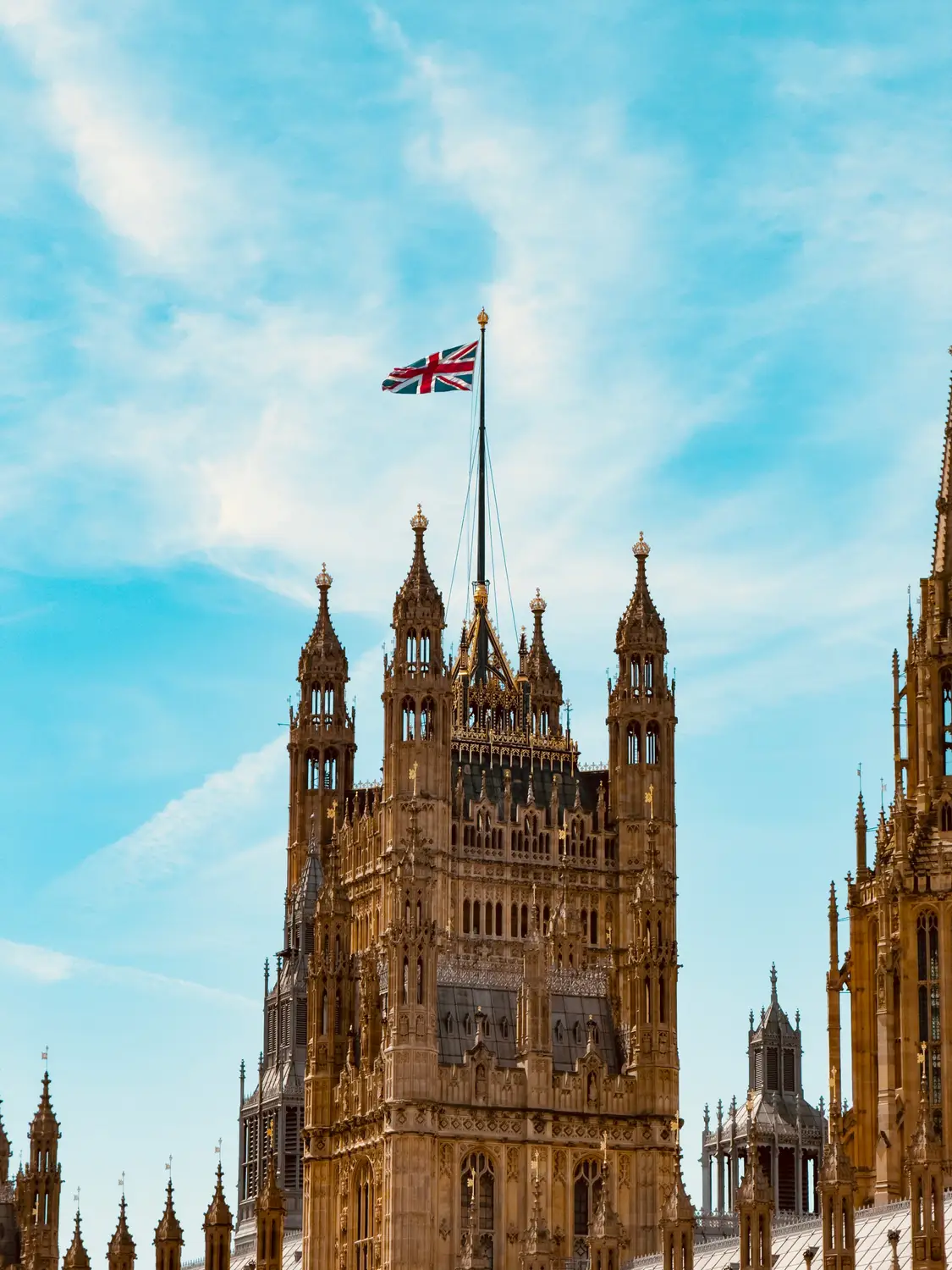 Ornate gothic pinnacles of the Houses of Parliament flying the Union Jack against a bright turquoise sky.