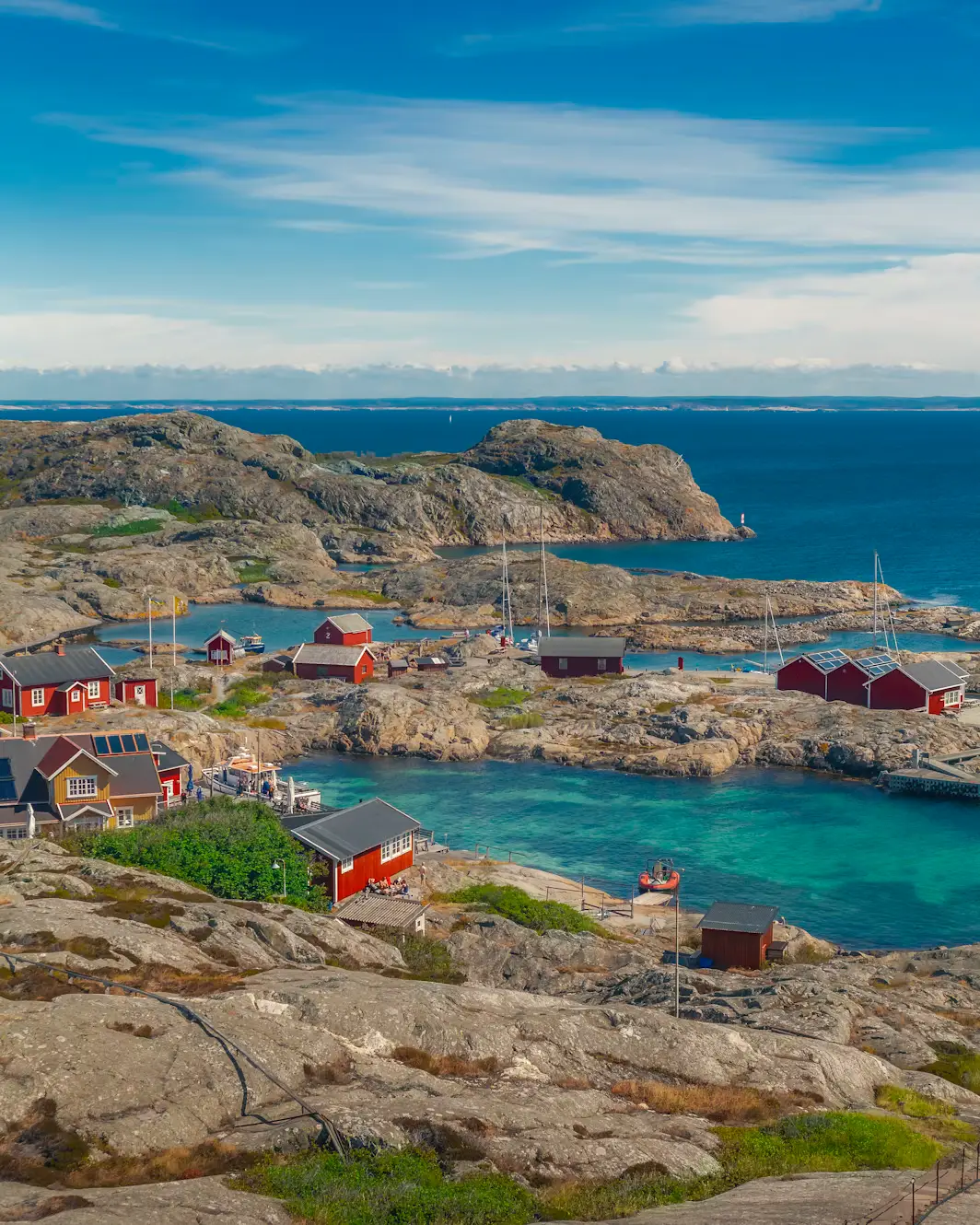 A small coastal fishing village with red and white houses nestled among rocky, weathered shoreline, overlooking a calm blue harbor.
