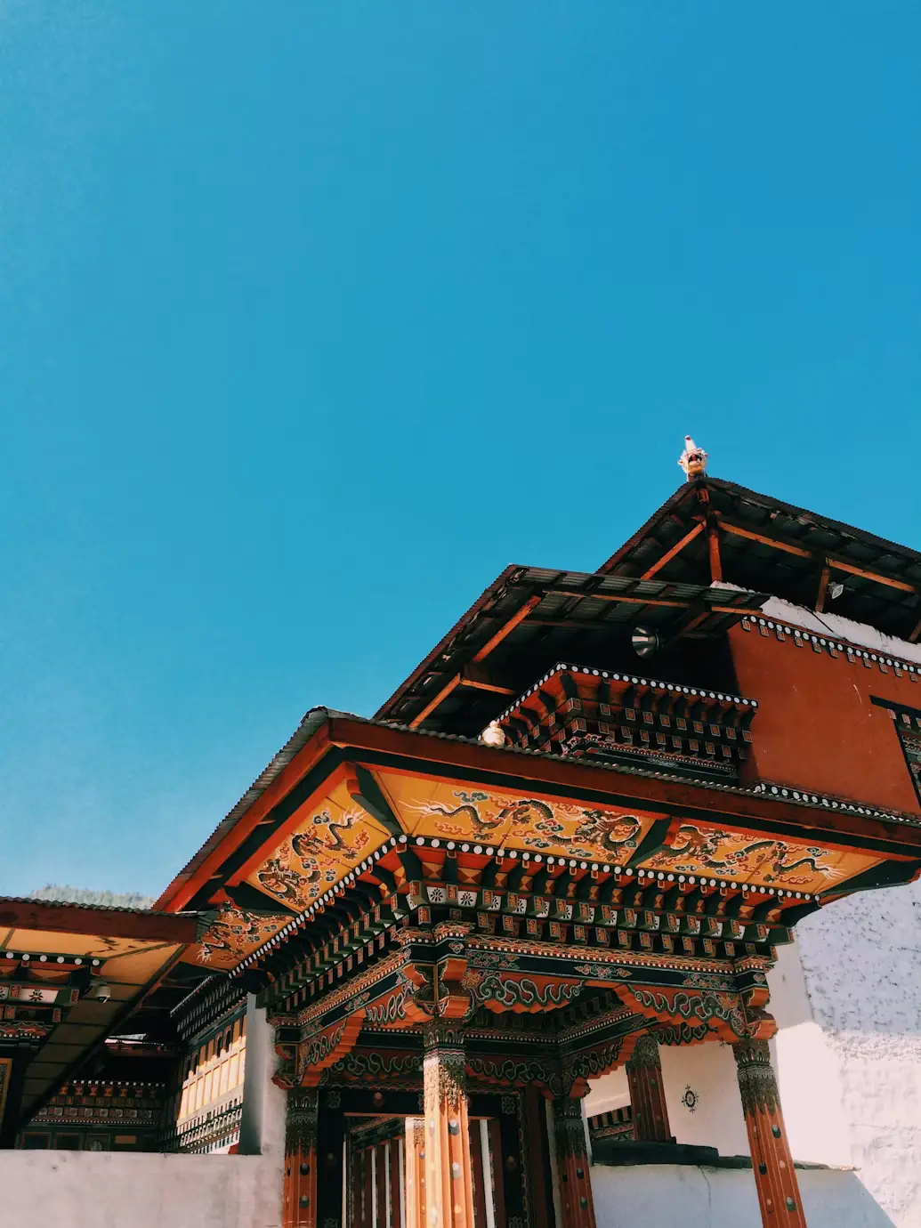 Low-angle view of the ornately carved wooden eaves and tiered roof of a traditional Bhutanese temple against a clear blue sky in Bhutan.