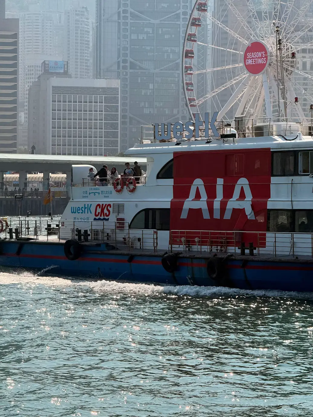 Star Ferry boat with AIA branding crossing Victoria Harbour past the Hong Kong Observation Wheel in Hong Kong.