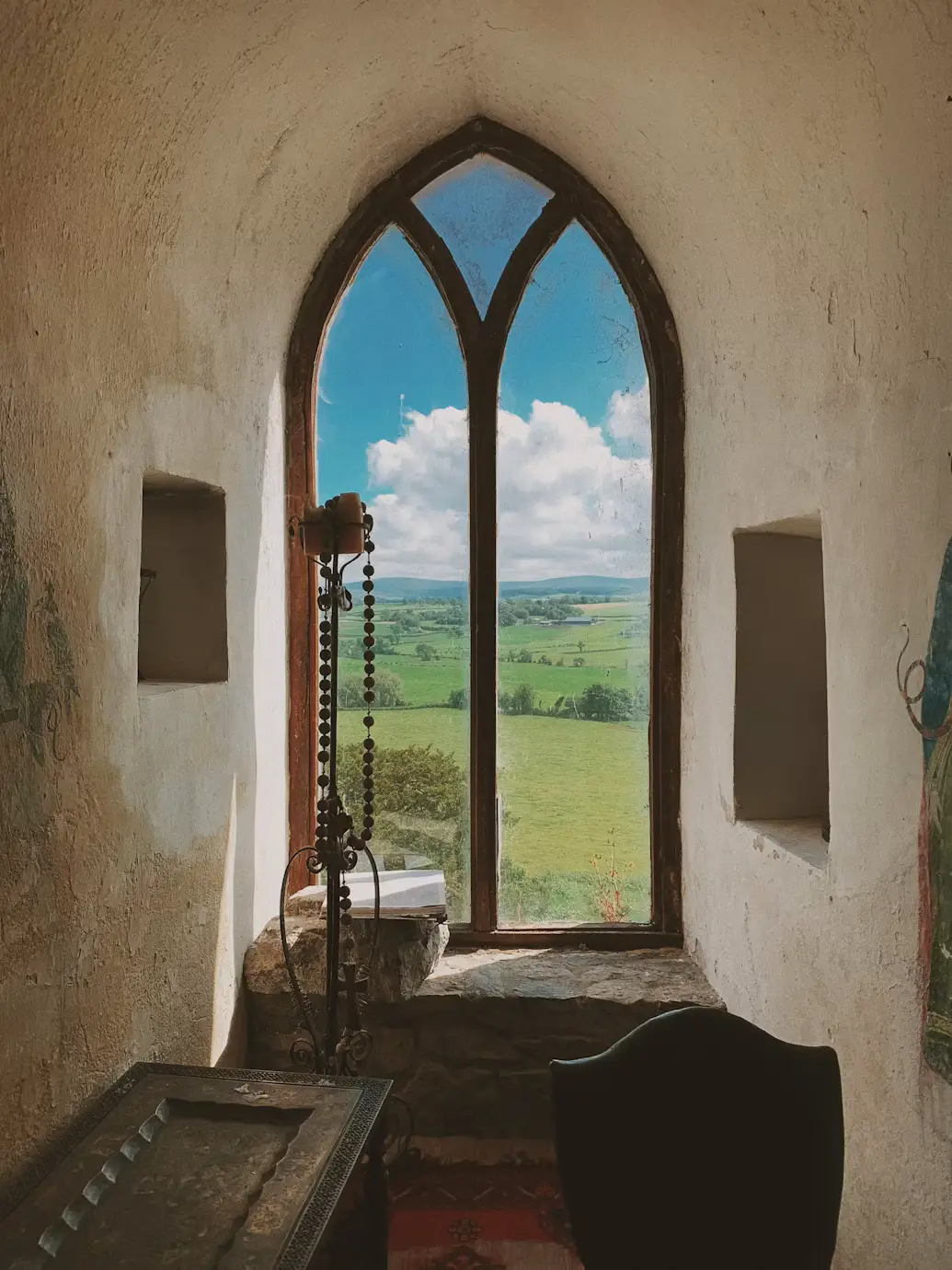 Gothic arched window inside a stone tower, framing a view of green countryside.