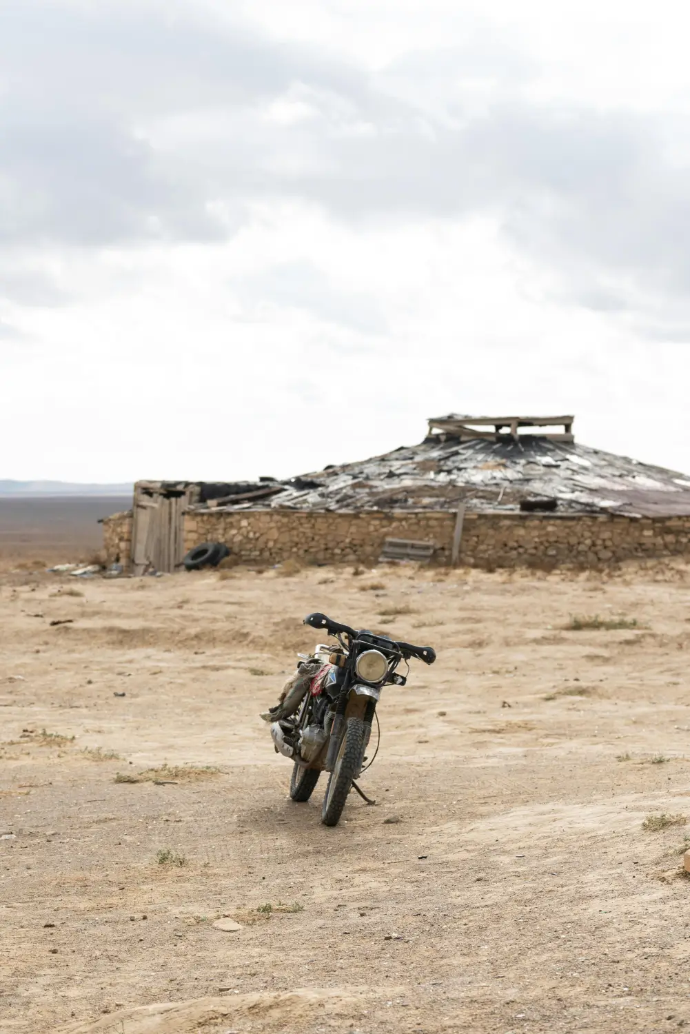 Vintage Soviet-era motorcycle parked on sandy desert ground in front of a ruined stone and wood structure in the Gobi Desert, Mongolia
