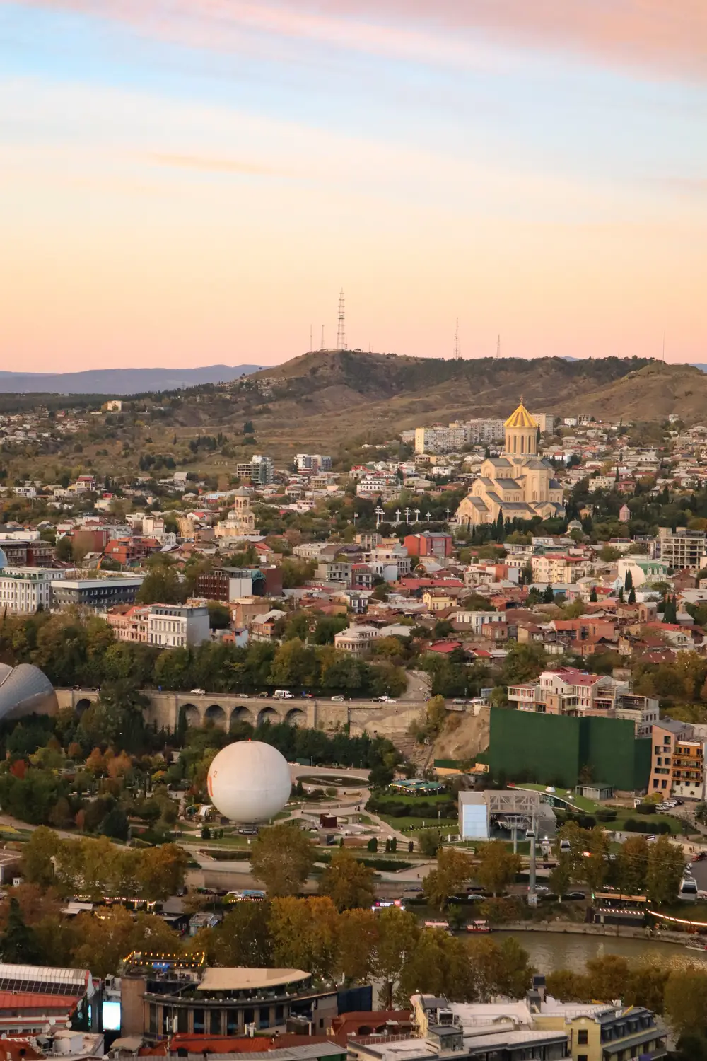 A cityscape with a large cathedral, hillside neighborhoods, a river, and a domed park structure beneath a pastel sunset sky.