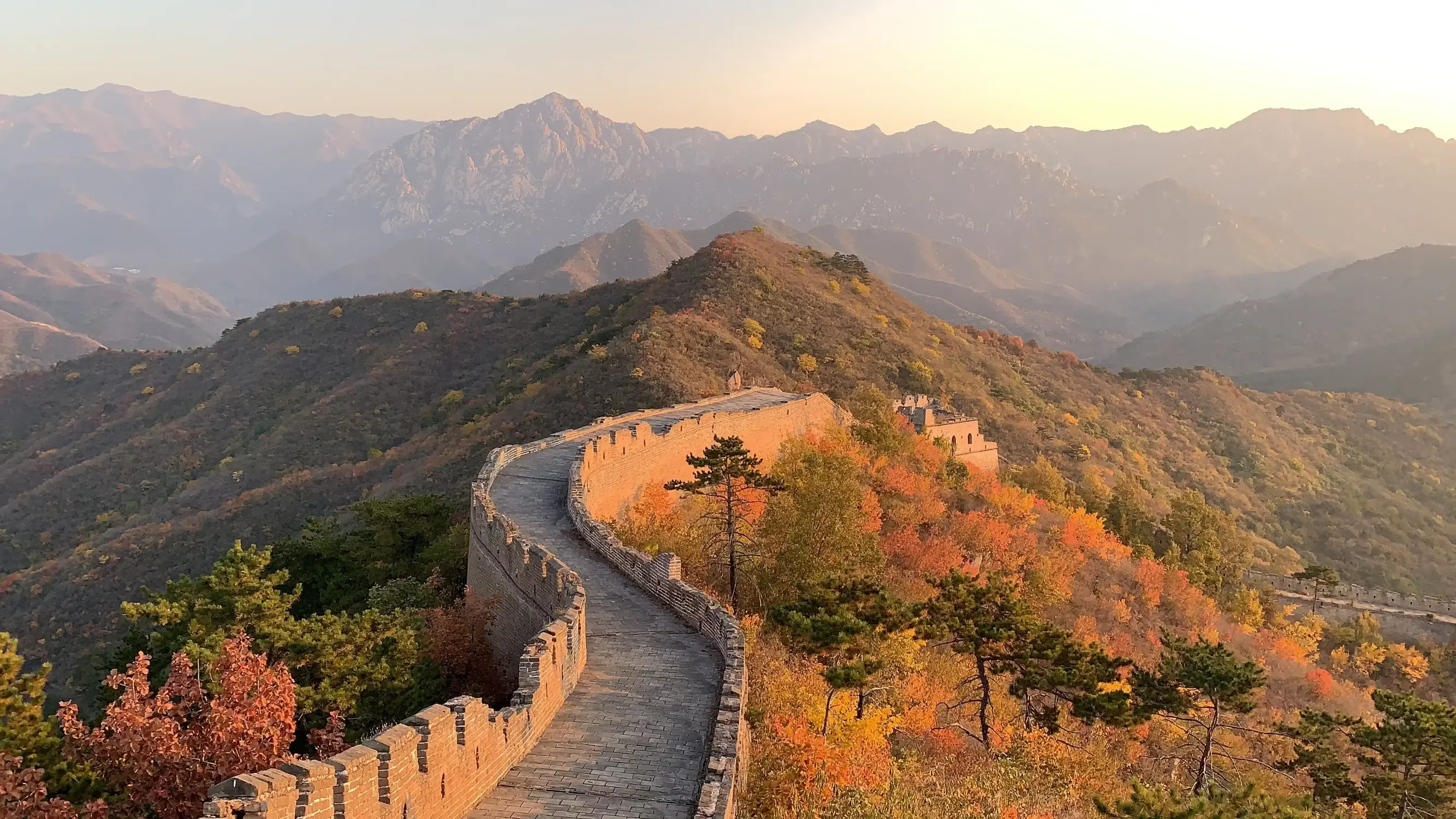 Great Wall of China winding through autumn mountain landscape at golden hour sunset, with colorful fall foliage in red, orange and yellow, misty mountain ranges in background