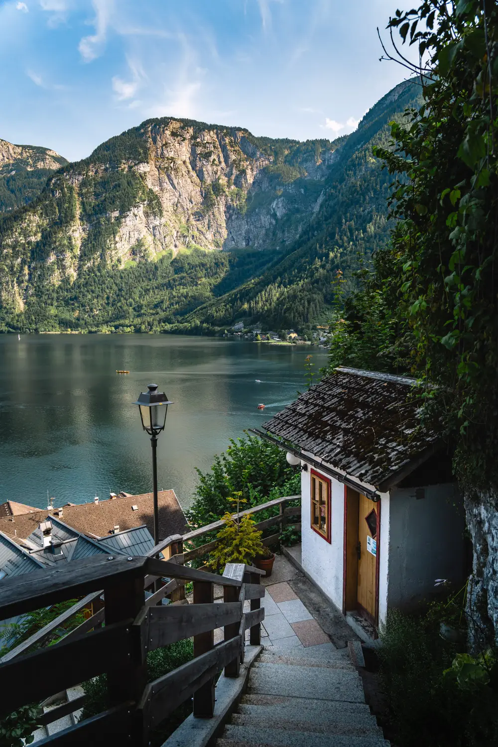 A quiet lakeside path with a small wooden building, leading toward a calm lake surrounded by steep, tree-covered mountains.