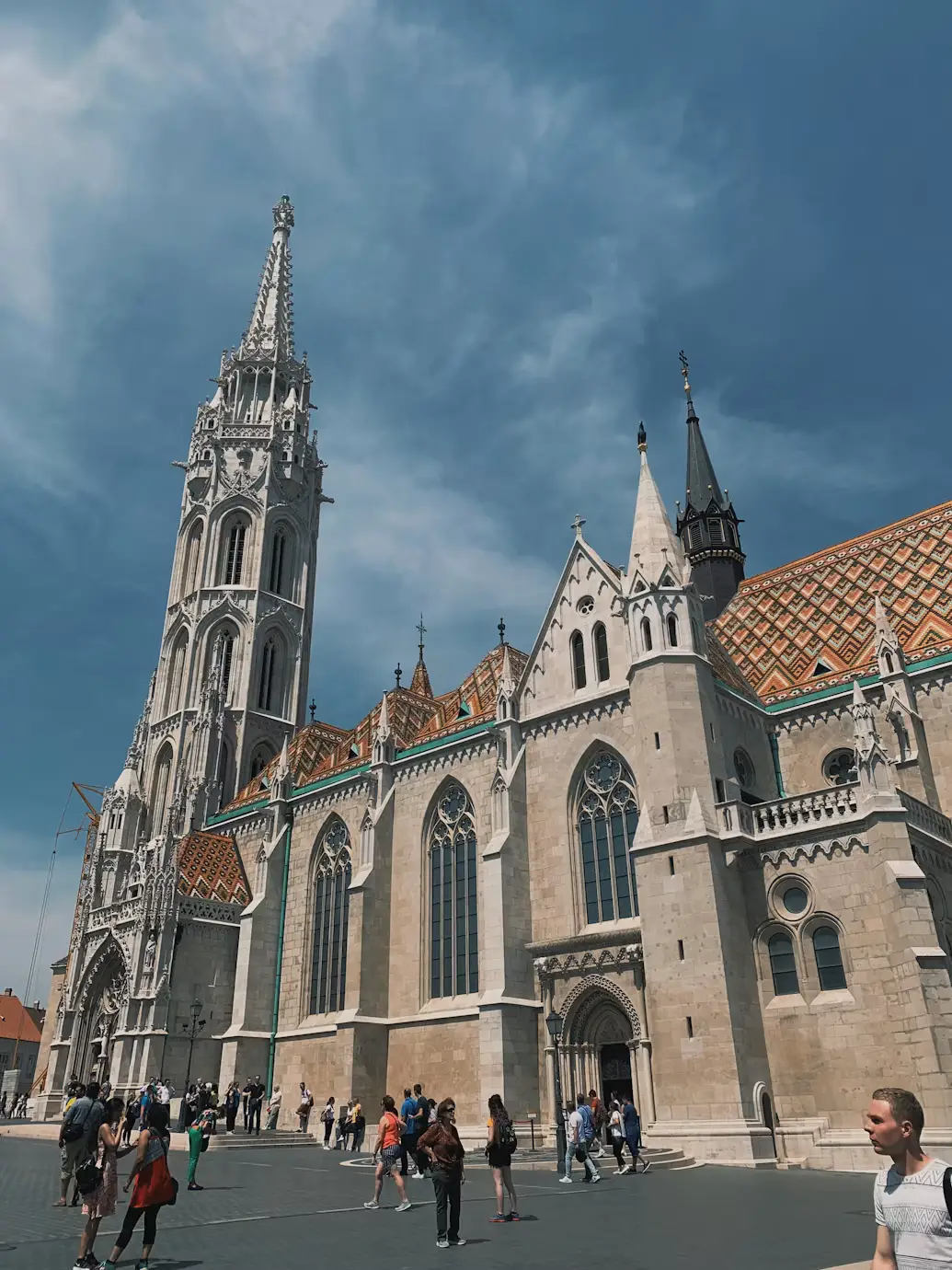 Gothic church with a tall ornate spire and patterned roof under a dramatic blue sky.