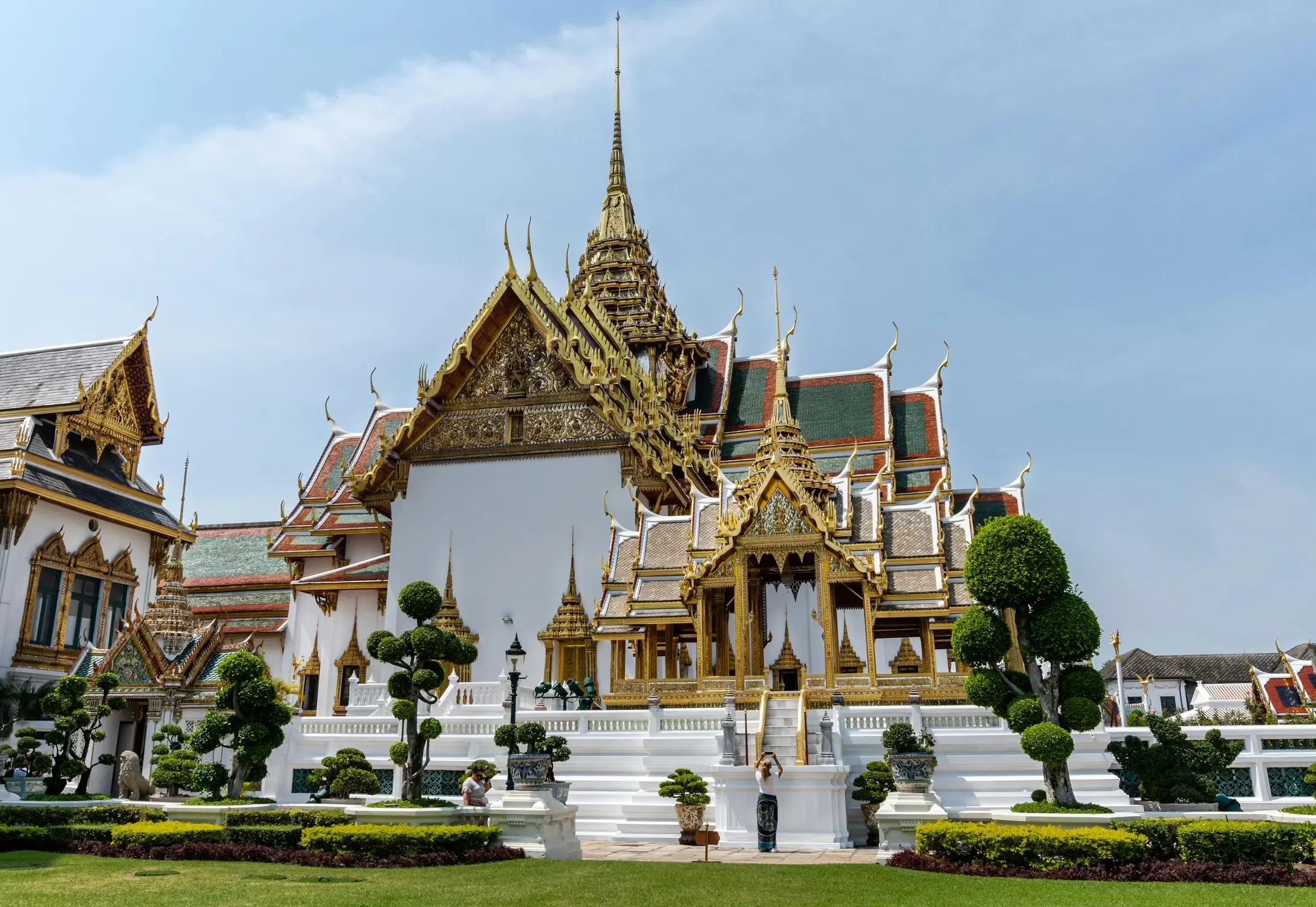 Phra Thinang Dusit Maha Prasat throne hall at the Grand Palace, Bangkok, with gilded multi-tiered roofs, golden spire, and manicured topiary gardens under a blue sky.