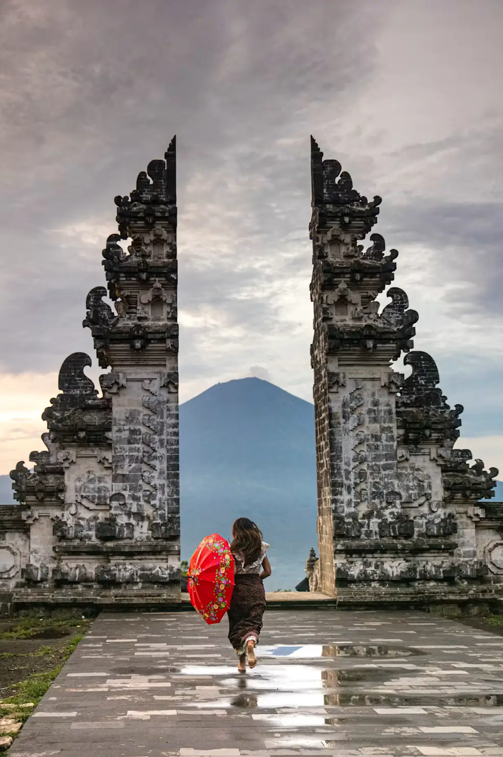 Person in traditional dress with a red umbrella standing at the Gates of Heaven (Lempuyang Temple) framing Mount Agung in Bali, Indonesia.