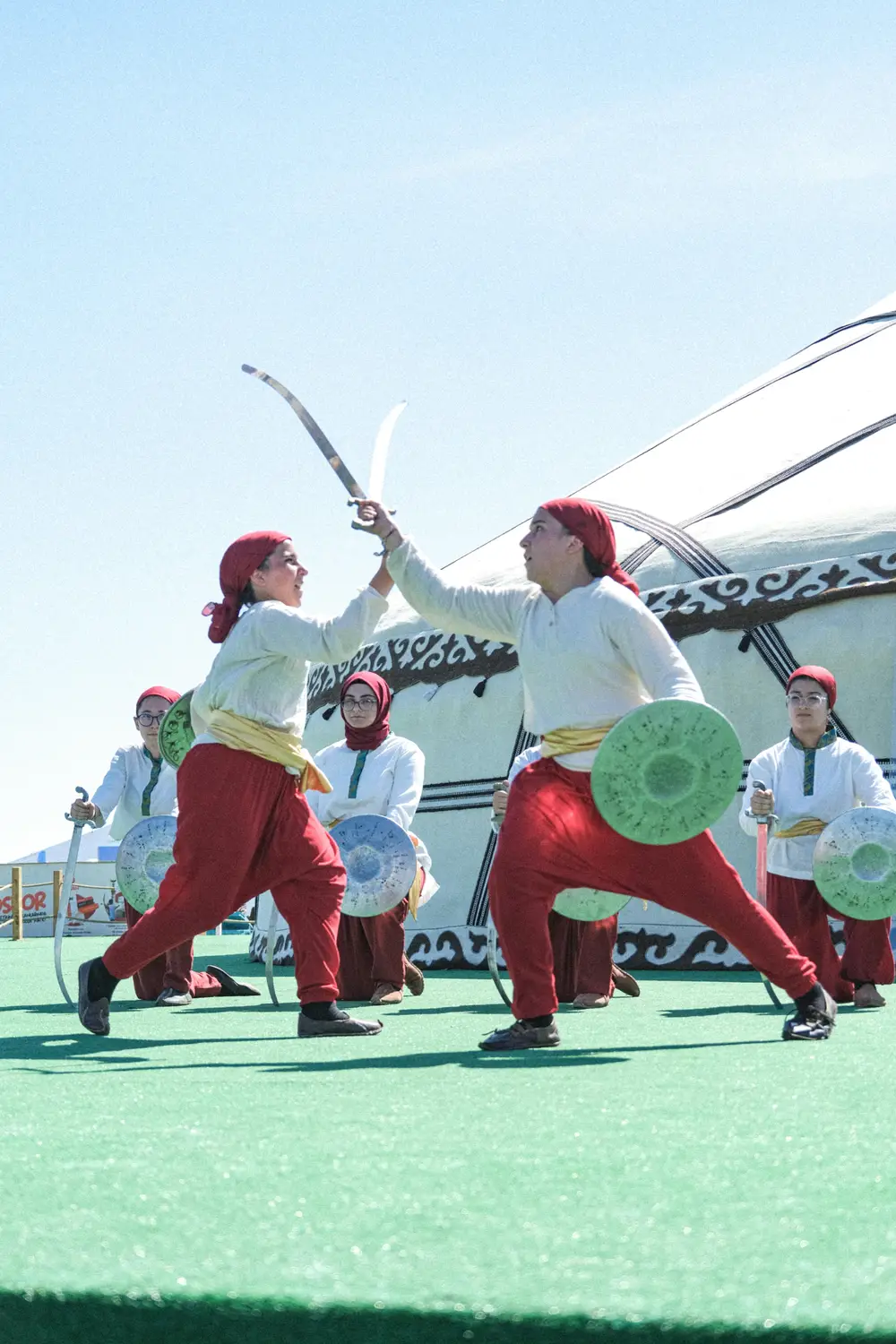 Performers in red trousers and white tunics clashing curved swords and round shields in a traditional martial display in front of a decorated yurt