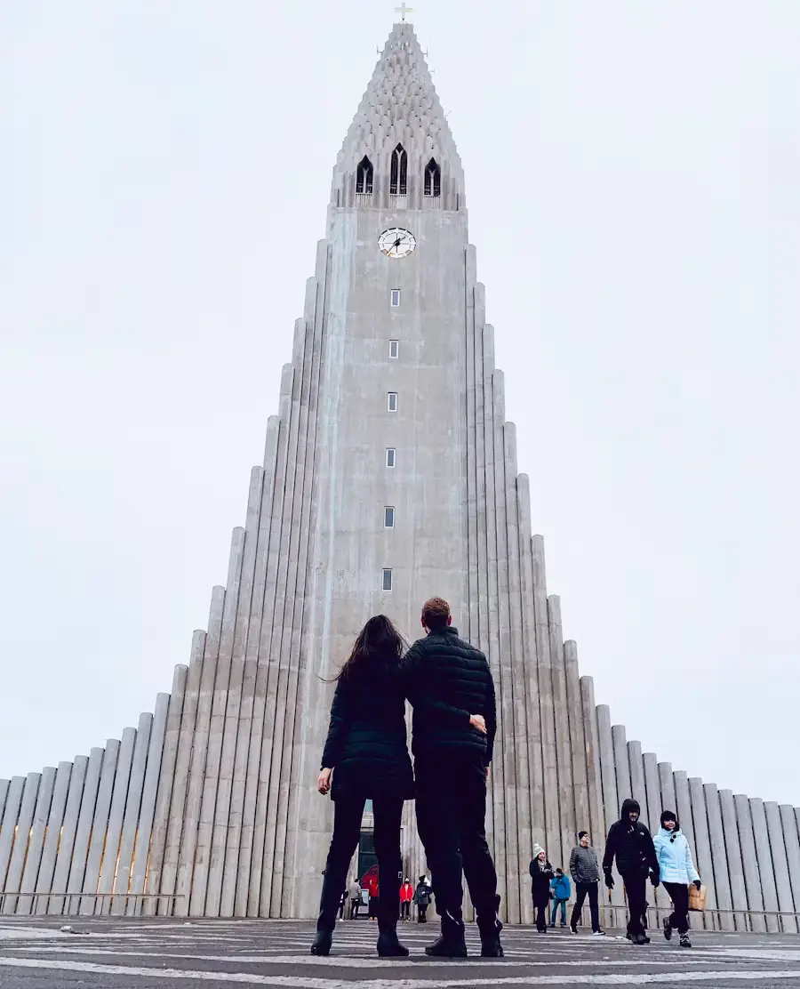Two people standing before the tall, organ-pipe-like facade of a ribbed concrete church tower, Iceland.