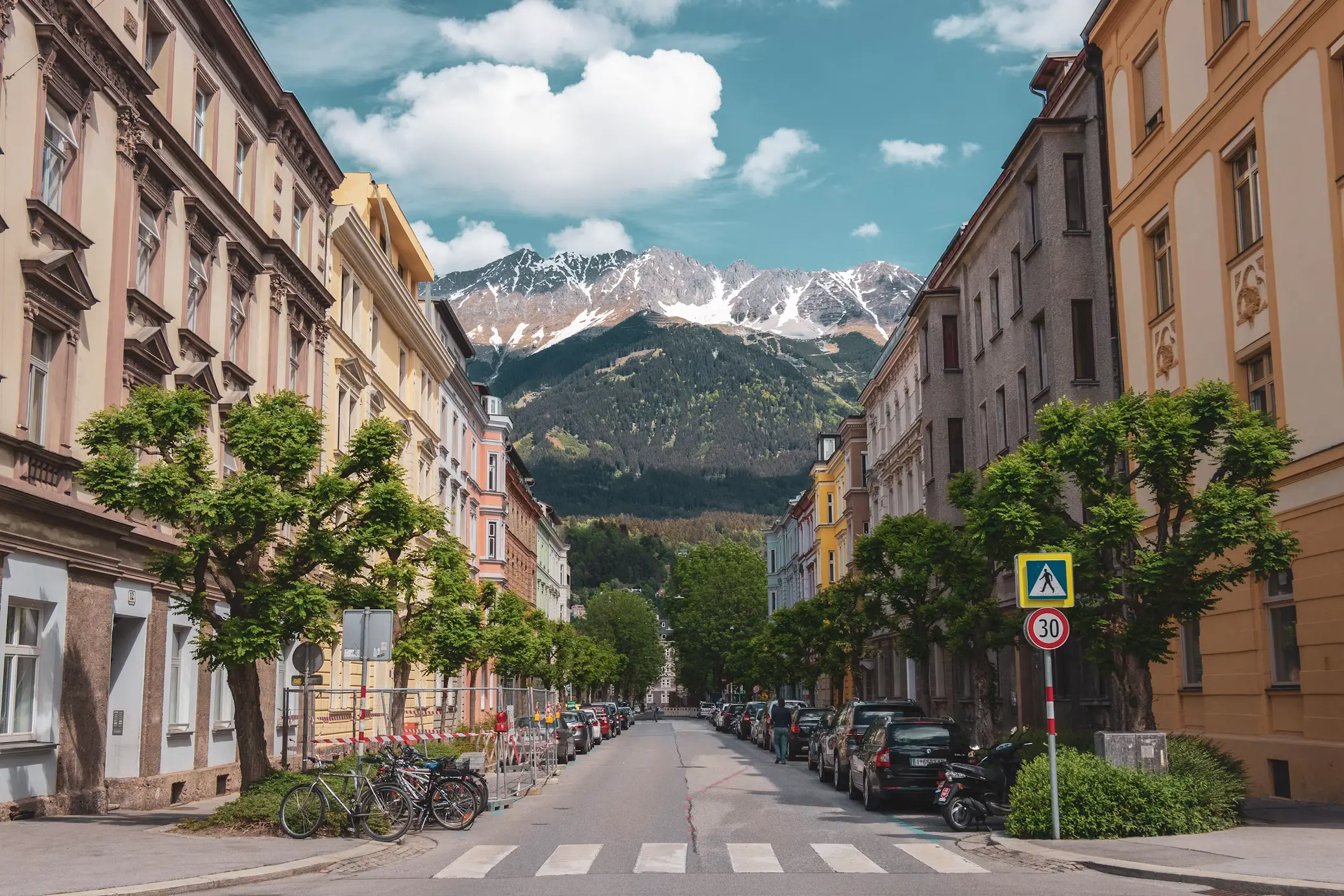A quiet cobblestone street lined with historic European apartment buildings, bicycles, and parked cars, framed by snow-capped mountains in the distance under a partly cloudy sky.