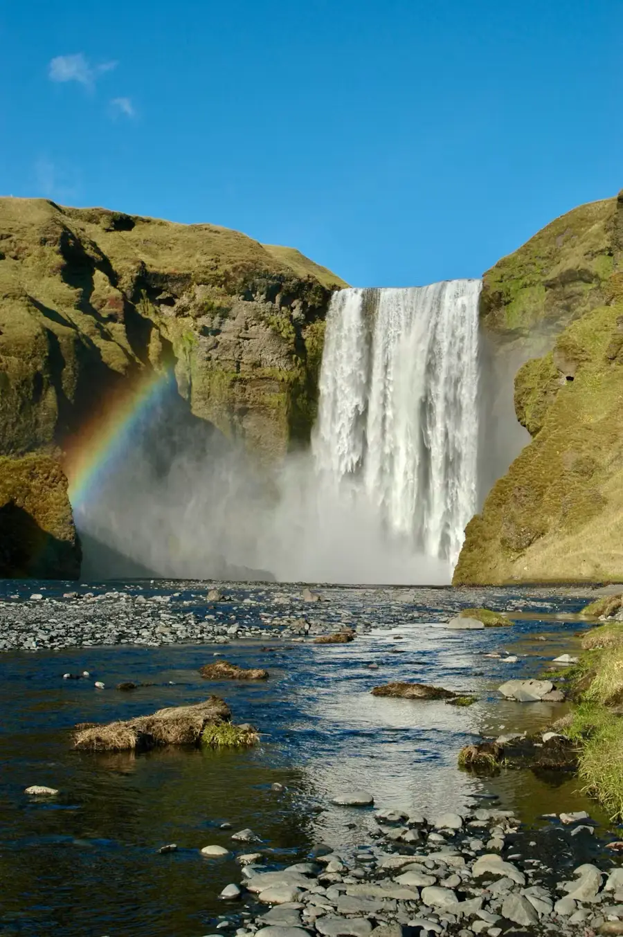 Waterfall pouring over a cliff into a rocky stream, with a rainbow forming in the mist, Iceland.