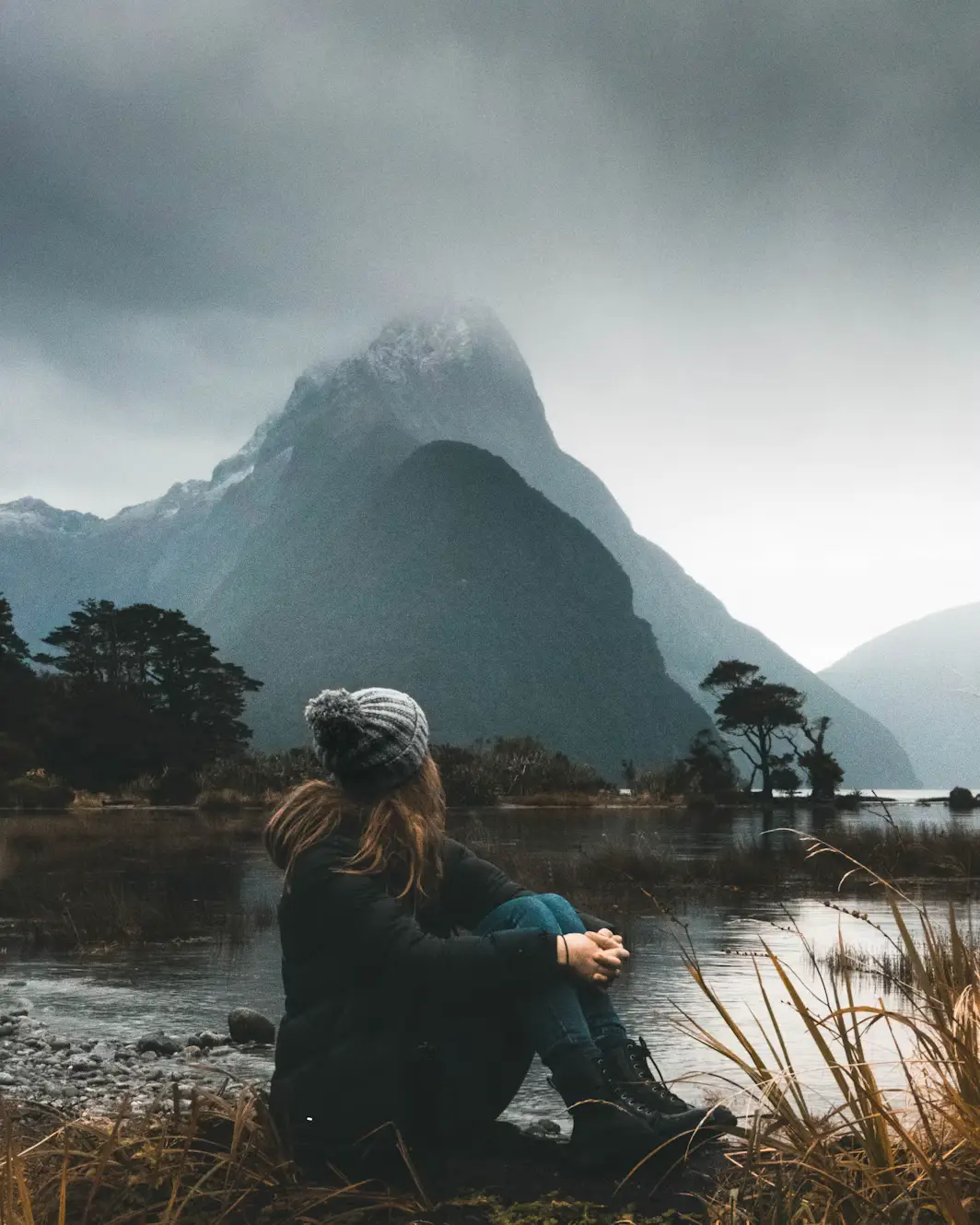 Person sitting by a calm lake facing a mist-covered mountain under dark clouds.