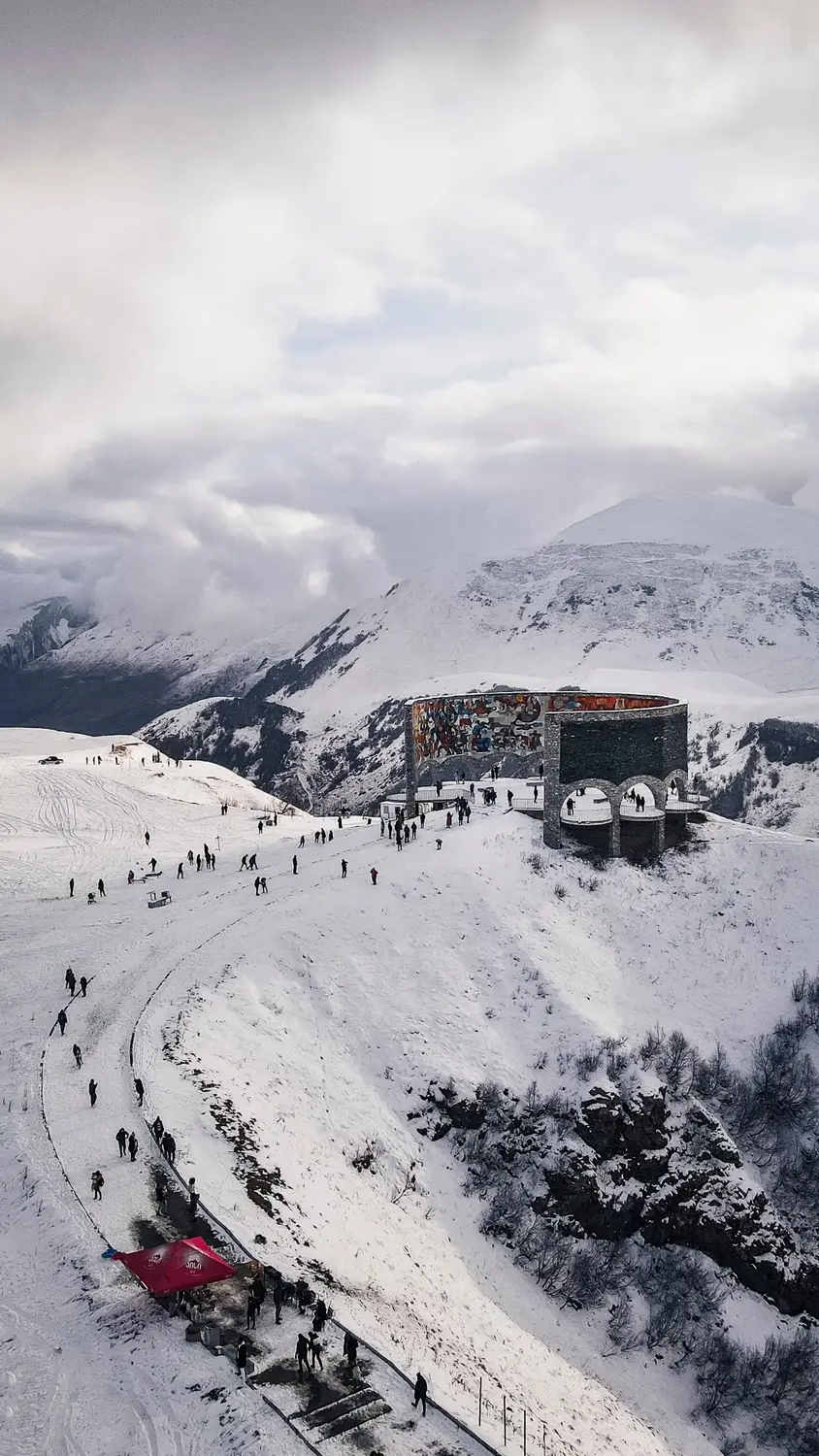 Snow-covered mountain slope with skiers and a colorful stone structure beneath cloudy peaks.