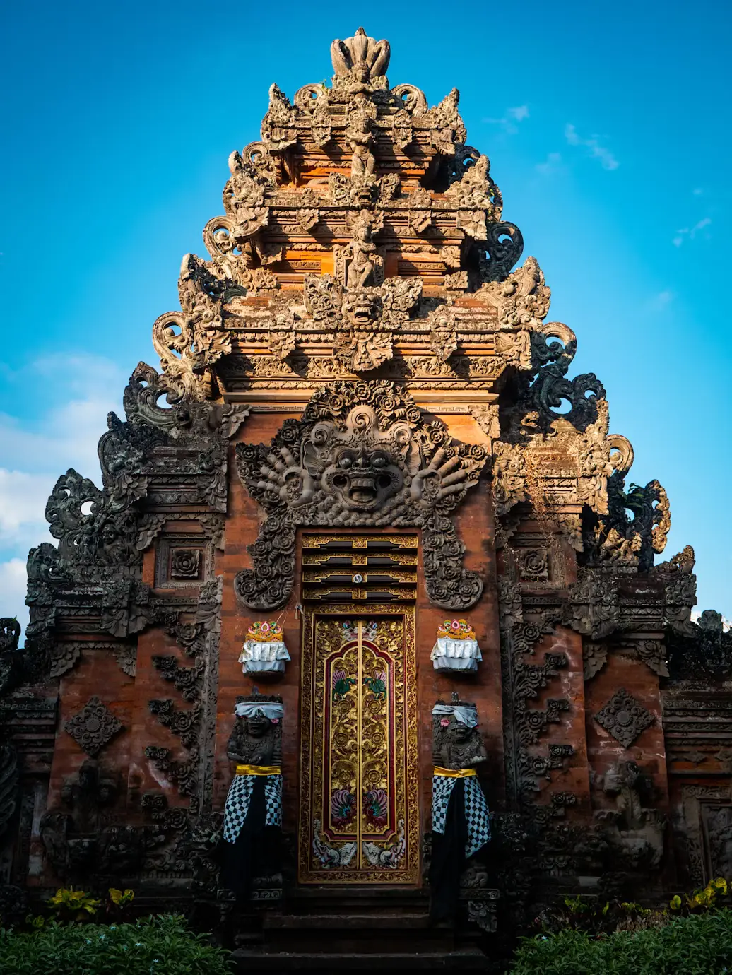 Intricately carved Balinese temple gate with a person in traditional dress holding a red umbrella against a blue sky in Bali, Indonesia.