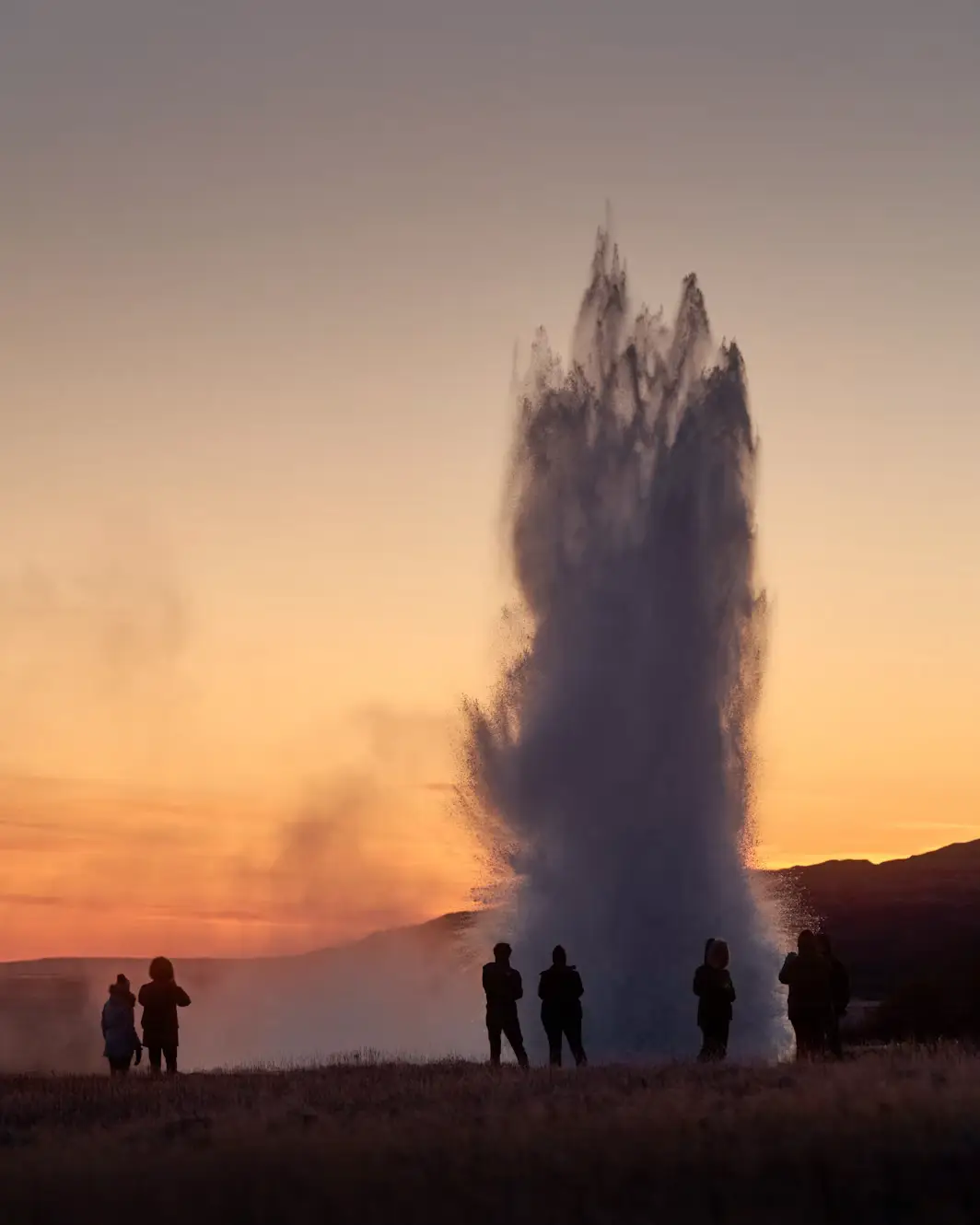 Silhouettes of people watching a geyser erupt against an orange sunset sky, Iceland.