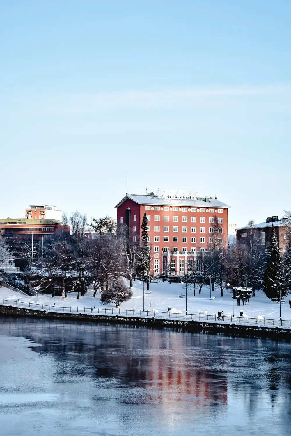 Snow-covered red building beside a calm river under a clear blue winter sky.