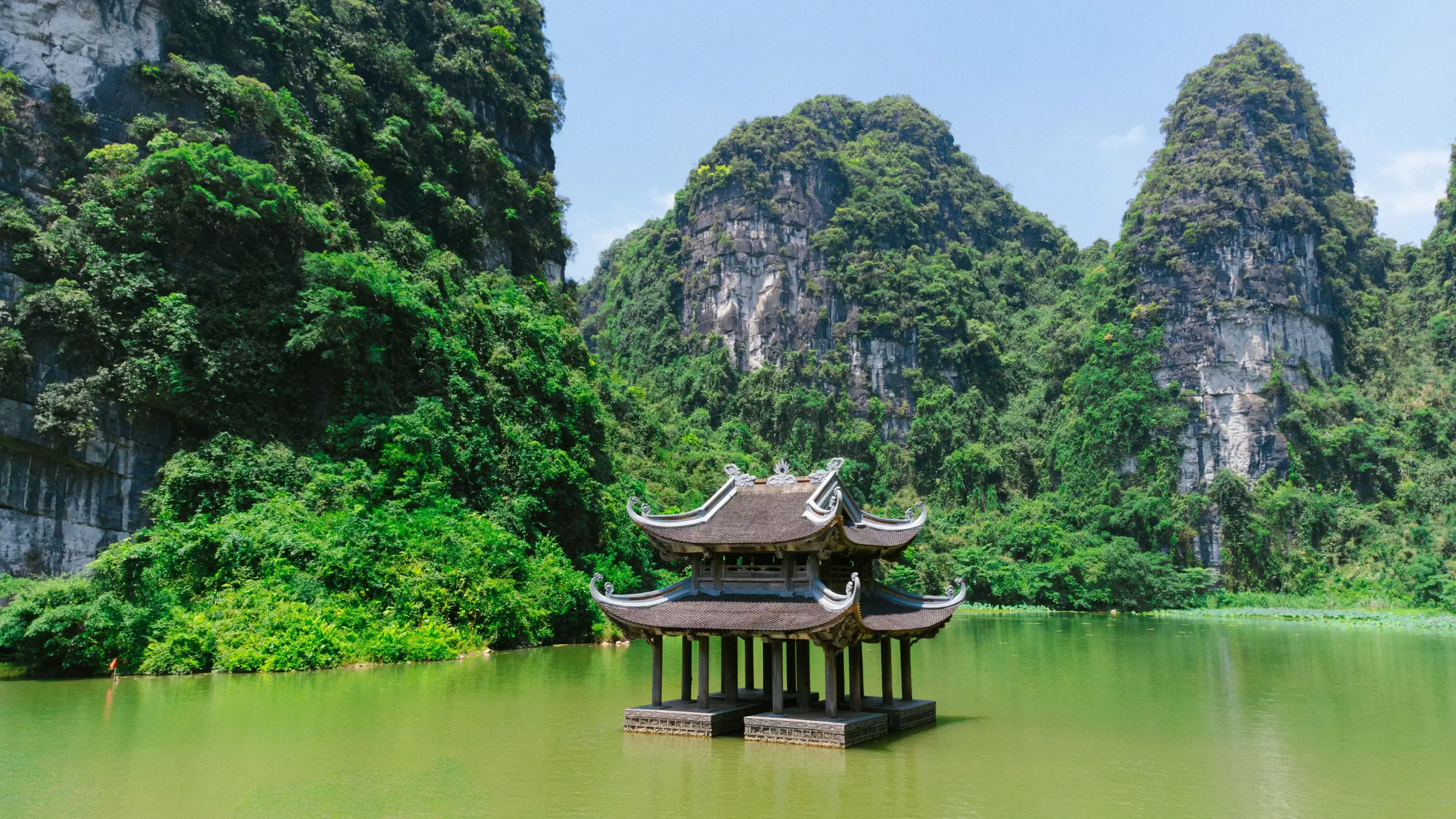 Traditional Vietnamese wooden pavilion with a curved tiled roof rising from calm green water, framed by towering limestone karst mountains covered in lush jungle in Trang An, Ninh Bình.