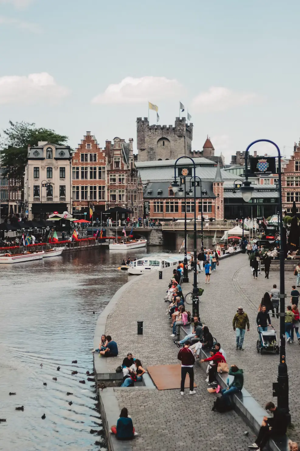 Ghent's Graslei waterfront with historic guild houses and the Gravensteen castle tower, crowds relaxing along the canal.