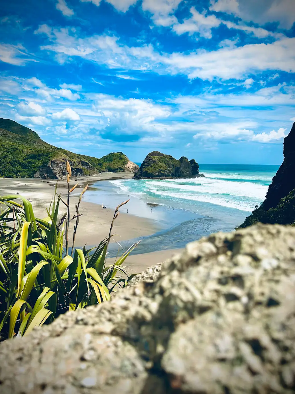 View of a sandy beach with turquoise water, rocky islets, and coastal vegetation.