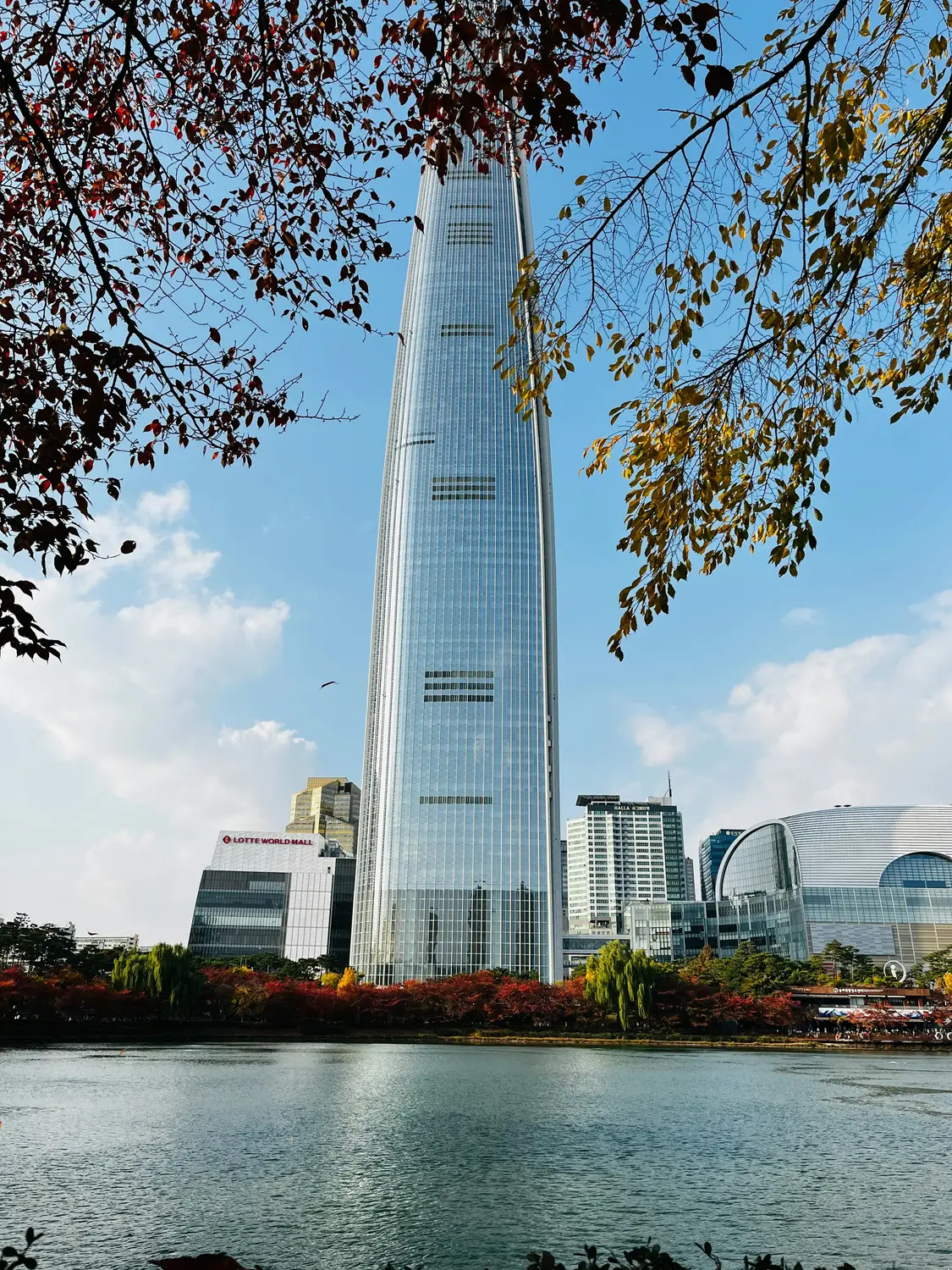 Lotte World Tower skyscraper rising above a lake and park with autumn trees in Seoul, South Korea.