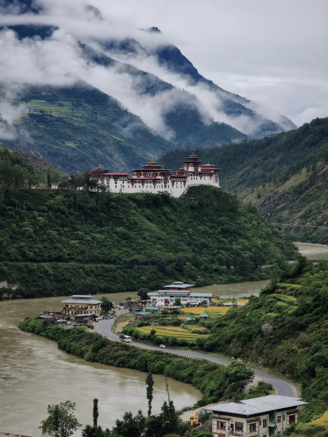 Traditional Bhutanese dzong perched on a green hillside above a winding river, framed by cloud-covered mountains in Bhutan.