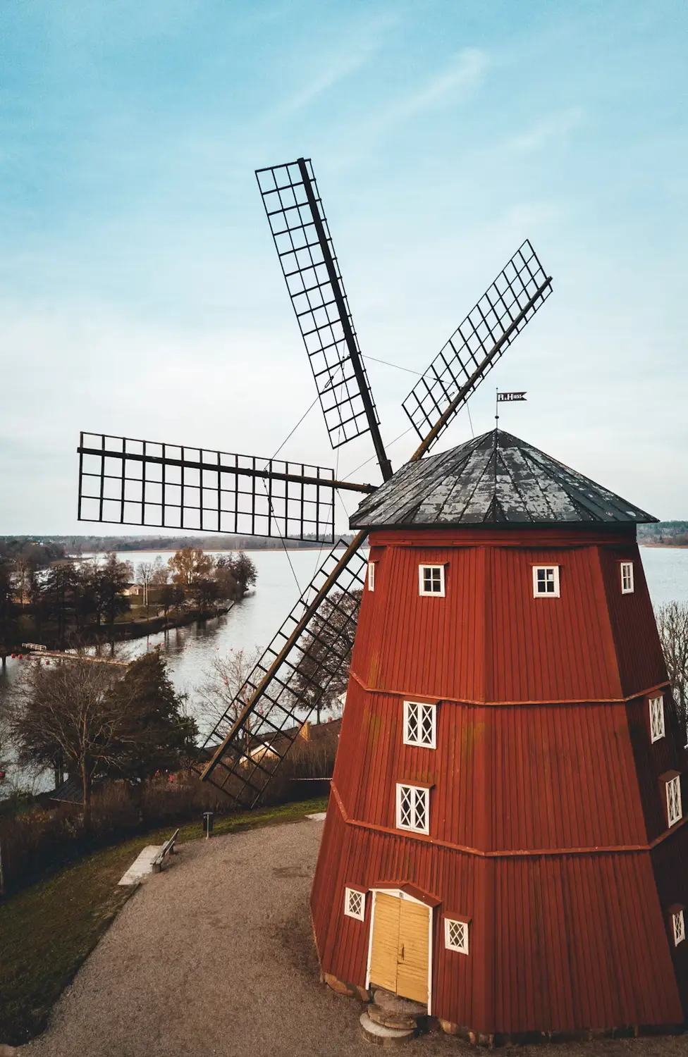 A traditional red wooden windmill with tall wooden sails stands beside a path near water, under a pale blue sky.