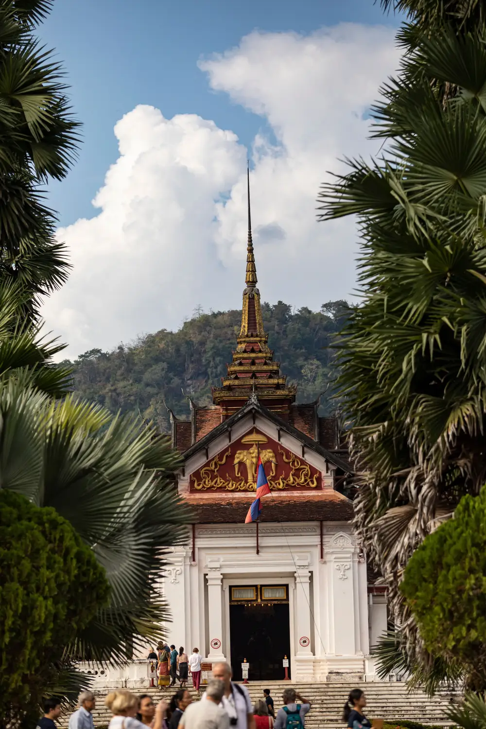 Gilded Buddhist temple with a tall spire framed by palm trees and a forested hillside in Laos.