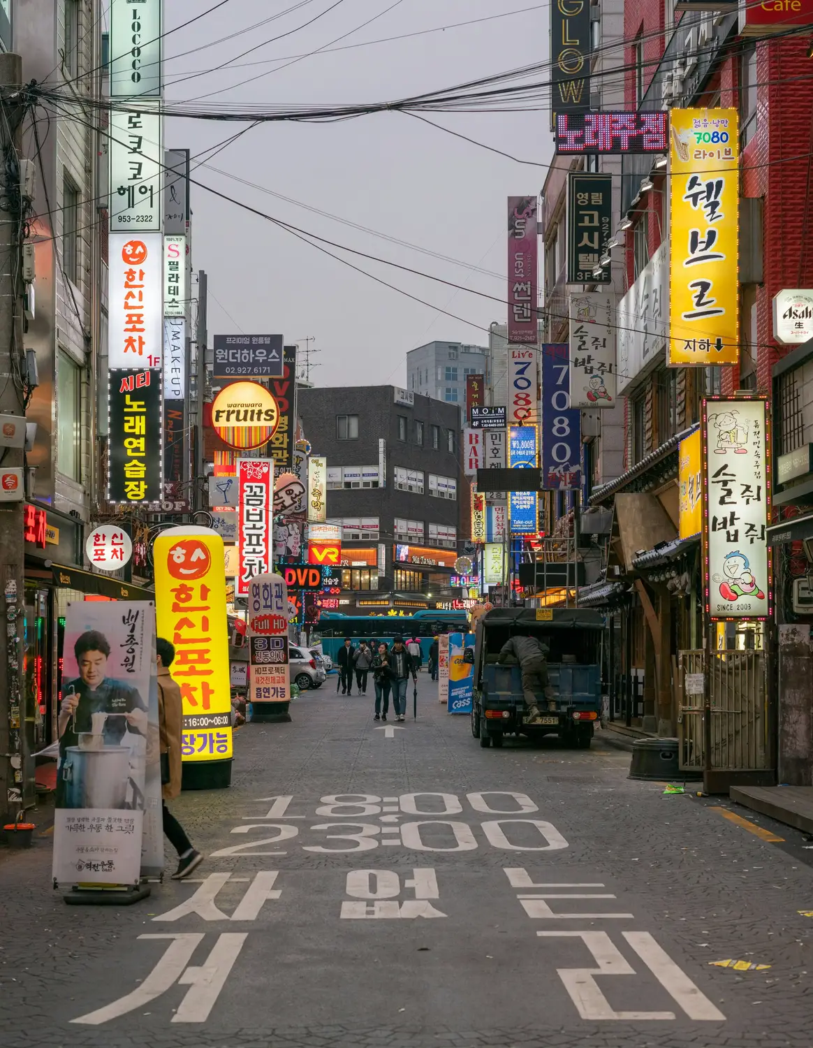 Narrow Korean street crowded with colorful Korean signage for bars, restaurants, and shops, with pedestrians and a delivery truck and Korean text painted on the road.